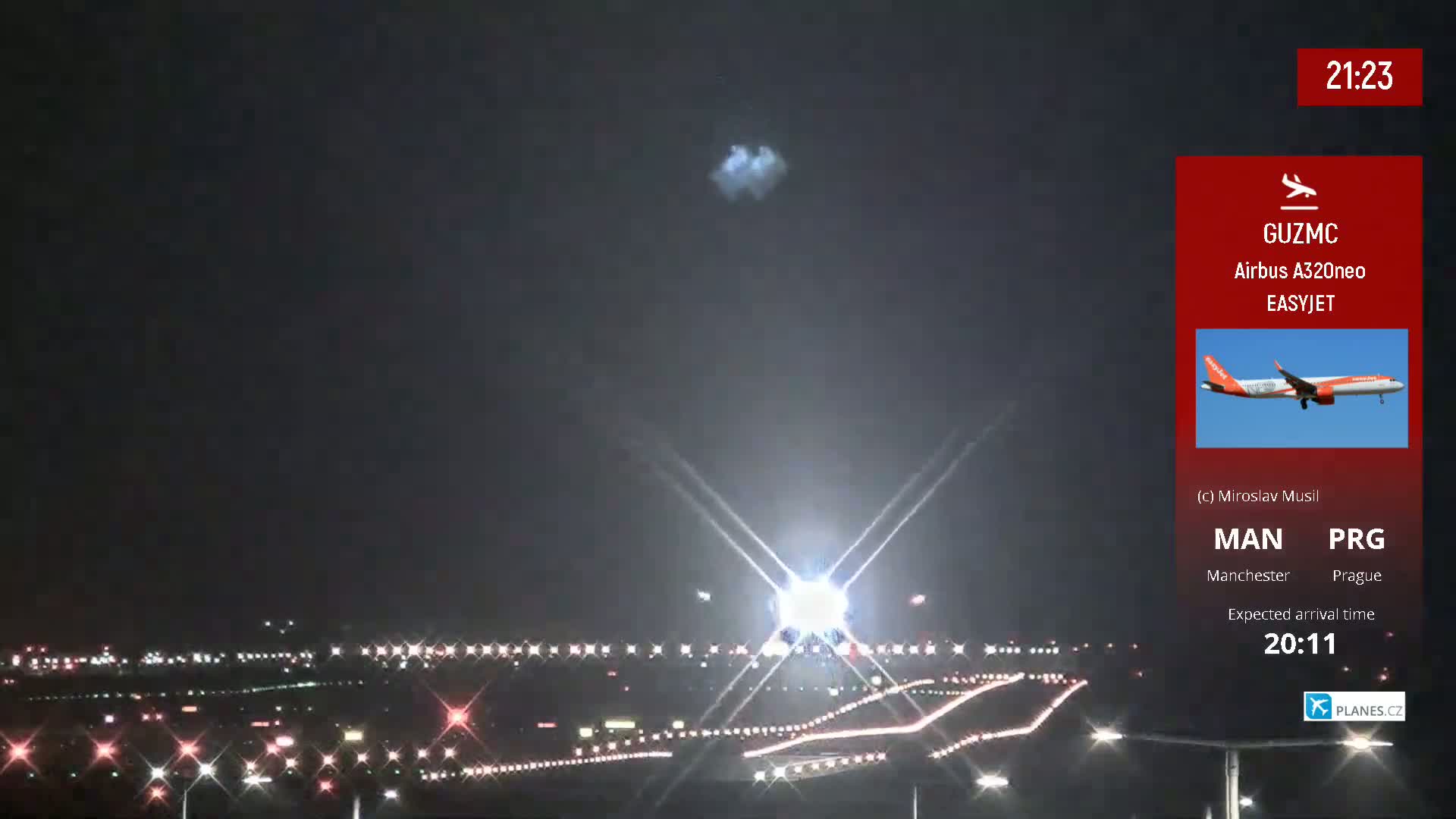 A nighttime long shot of an airport runway with lights, a bright light source in the center, and an airplane in the sky above.
