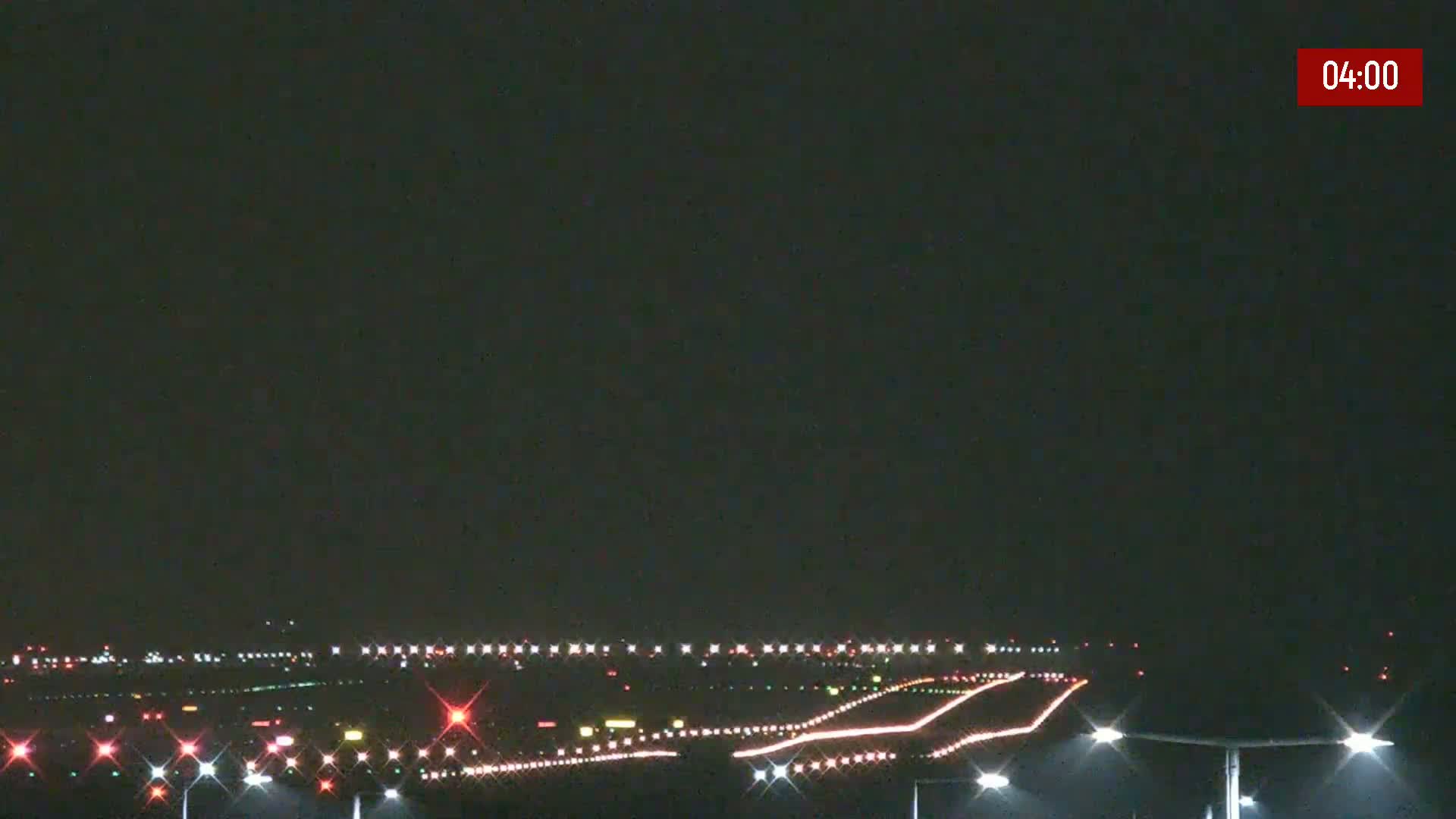 Under a dark night sky, the illuminated runways and taxiways of an airport are visible, with bright lights marking the ground.