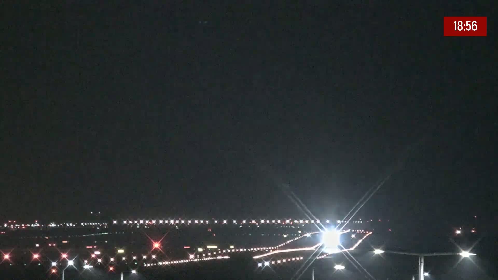 A nighttime view of an airport runway and taxiway with lights illuminating the ground and vehicles, under a clear, dark sky.