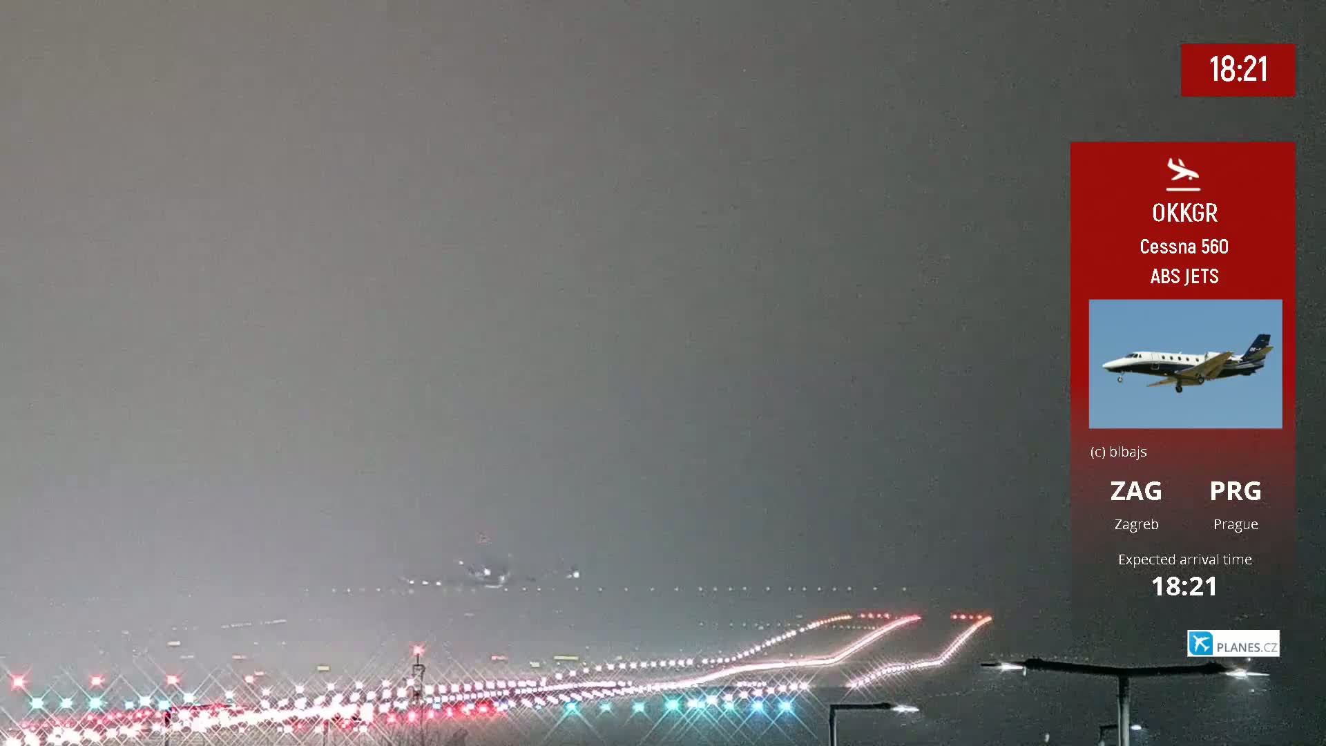 A long runway with brake lights from landing aircraft is visible under a hazy, gray sky at night.