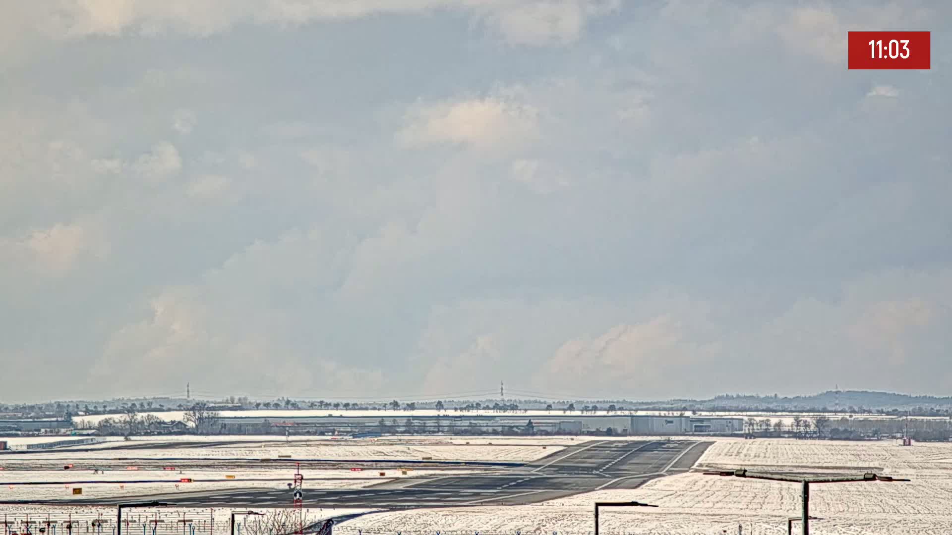 A snow-covered airport runway stretches toward a distant horizon under a mostly cloudy sky.