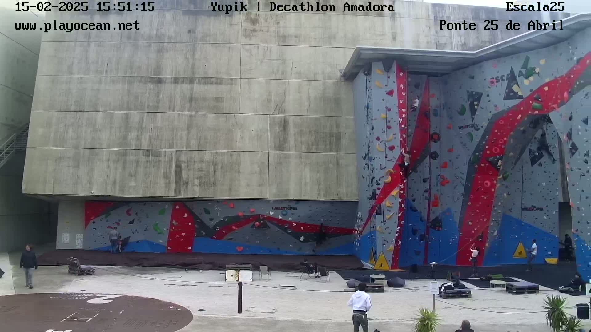 A large outdoor climbing wall with several people climbing and others sitting nearby on a cloudy day.