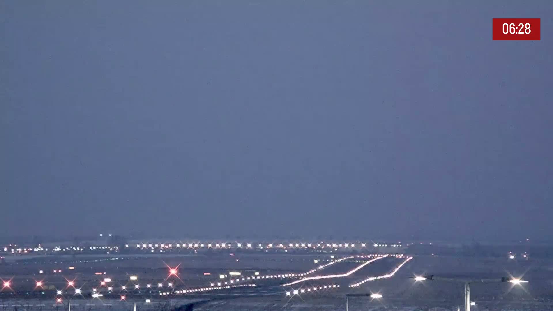 Under a dusky sky, a long stretch of highway with sparse traffic is illuminated by lights, leading towards a distant horizon dotted with more lights.