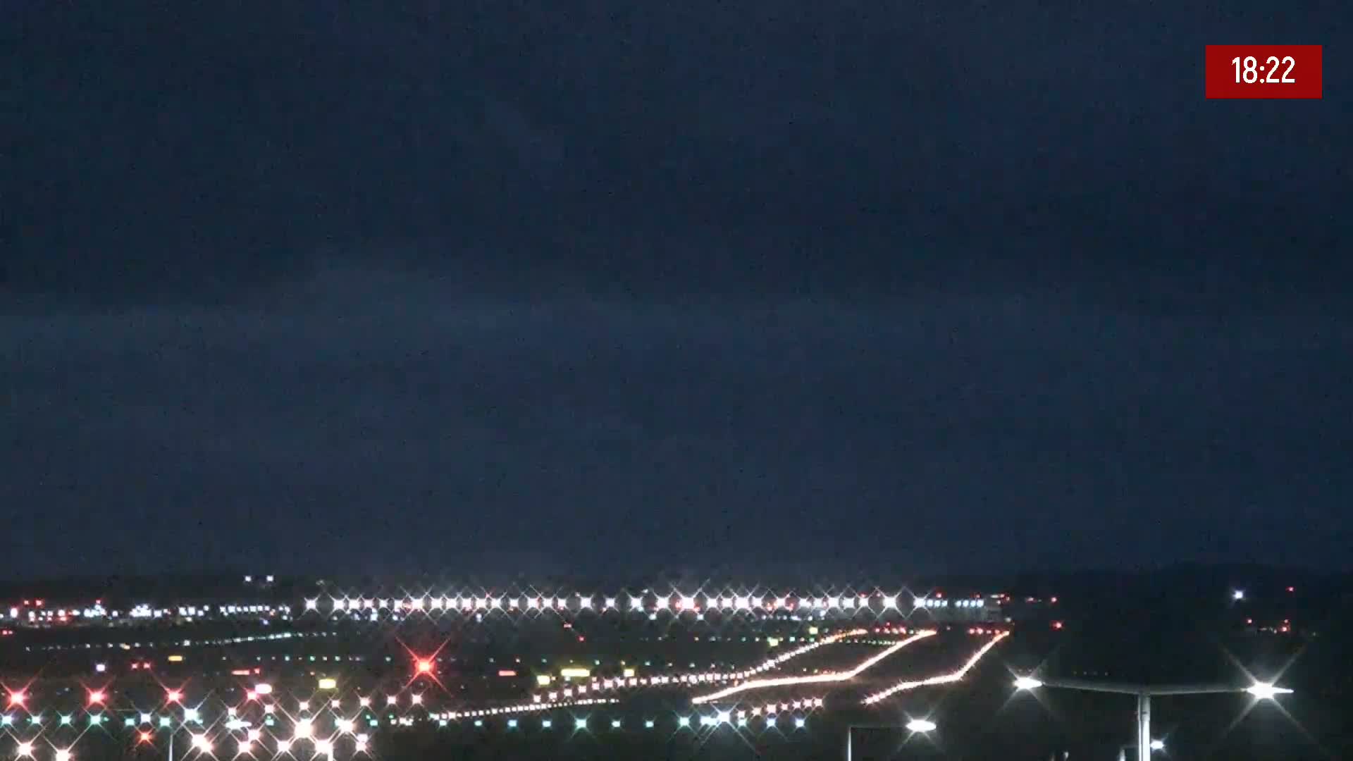 Under a dark night sky, a brightly lit airport runway shows various colored lights indicating taxiways and runways.