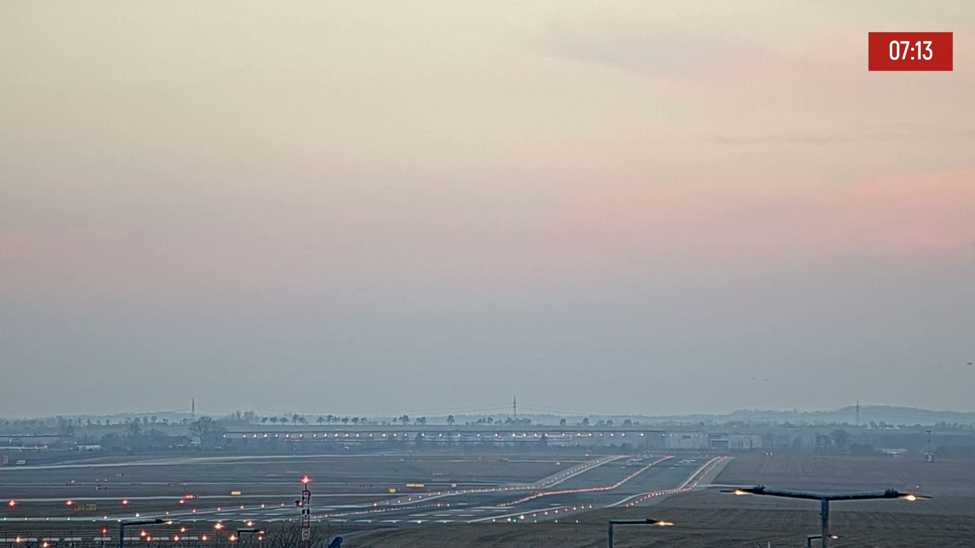 A hazy sunrise illuminates an airport runway and taxiways with lights, surrounded by flat land and distant trees.