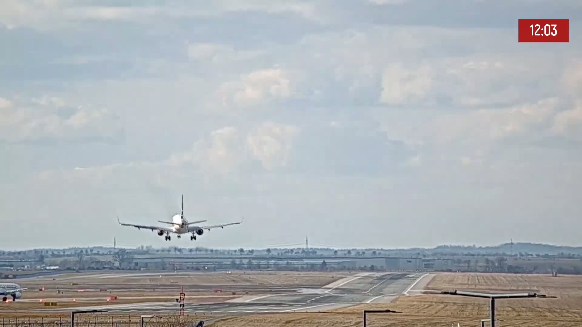 A commercial airplane is making a landing approach to an airport runway under a mostly cloudy sky.