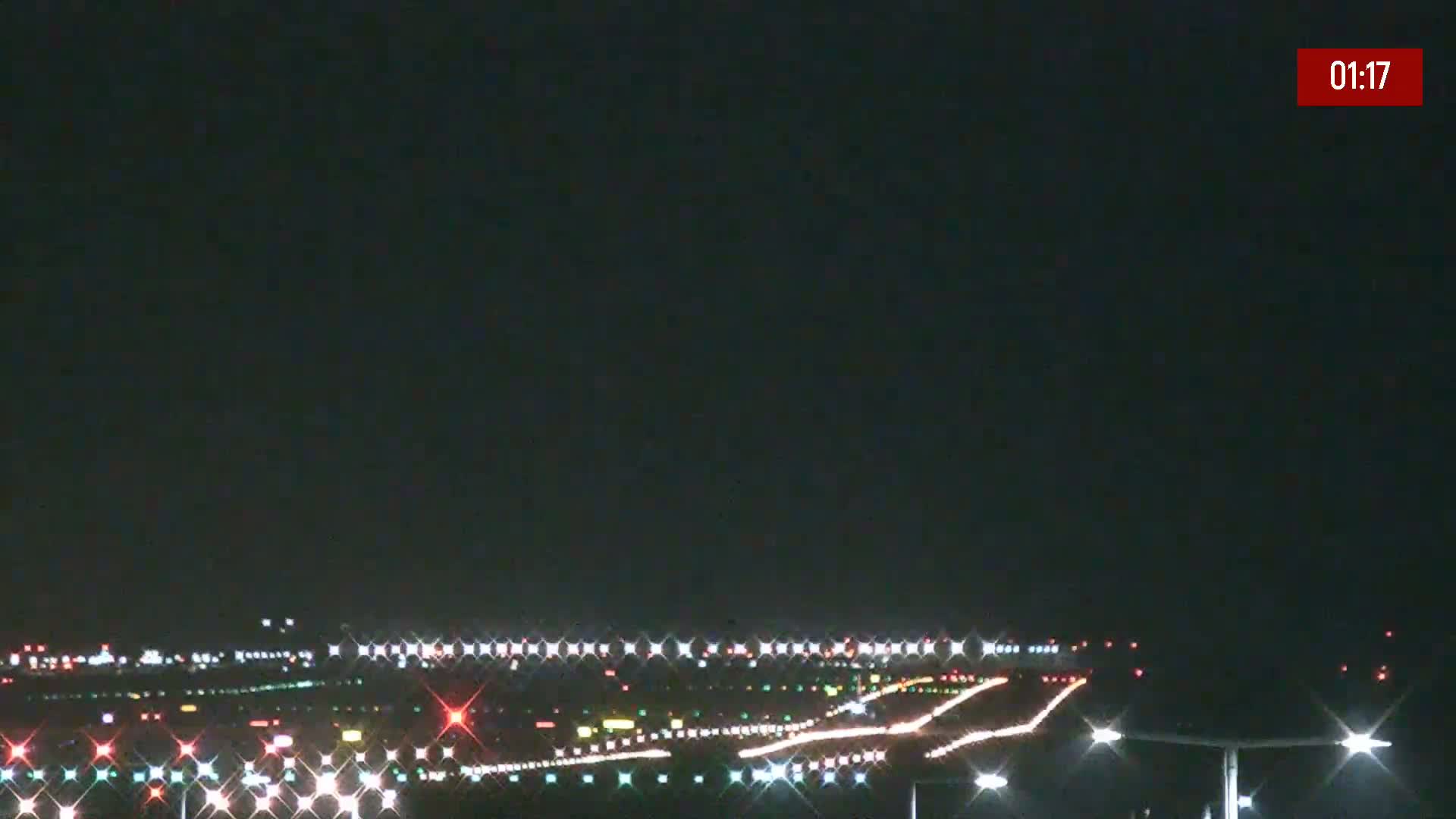At night, under a dark sky, airport runway lights illuminate the ground and a plane's lights streak across the tarmac.