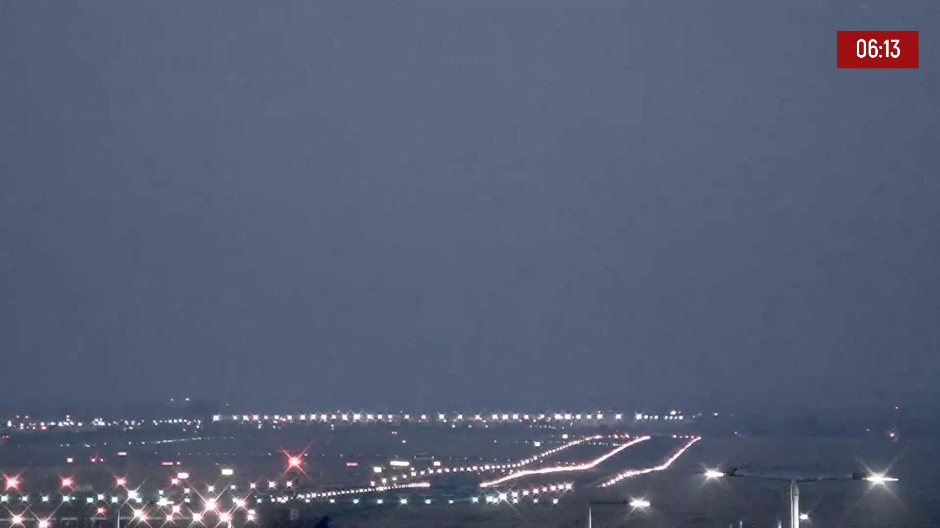 Under a dusky sky, the lights of numerous vehicles illuminate a long roadway and a distant city skyline.
