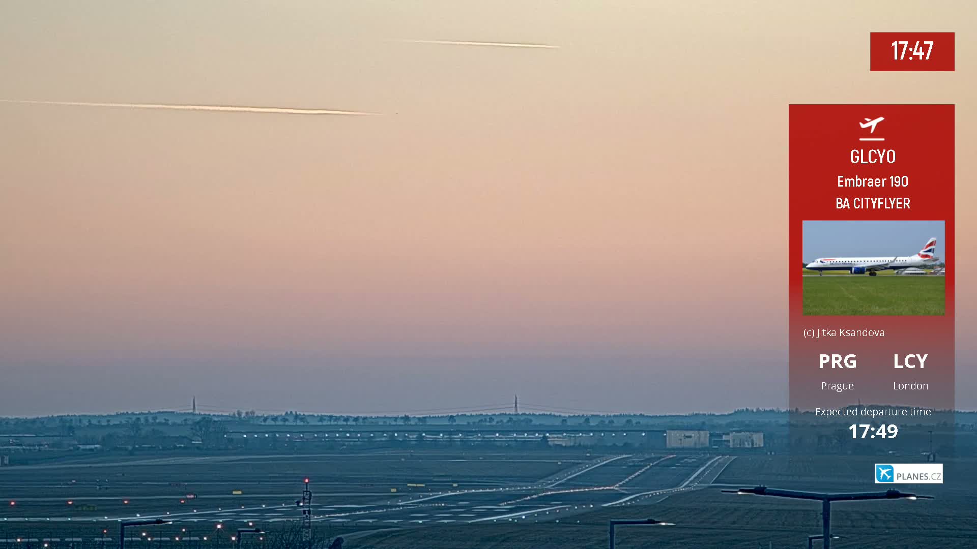A wide shot of an airport runway at dusk or dawn under a pale sky with contrails visible, showing taxiways and airfield lighting.