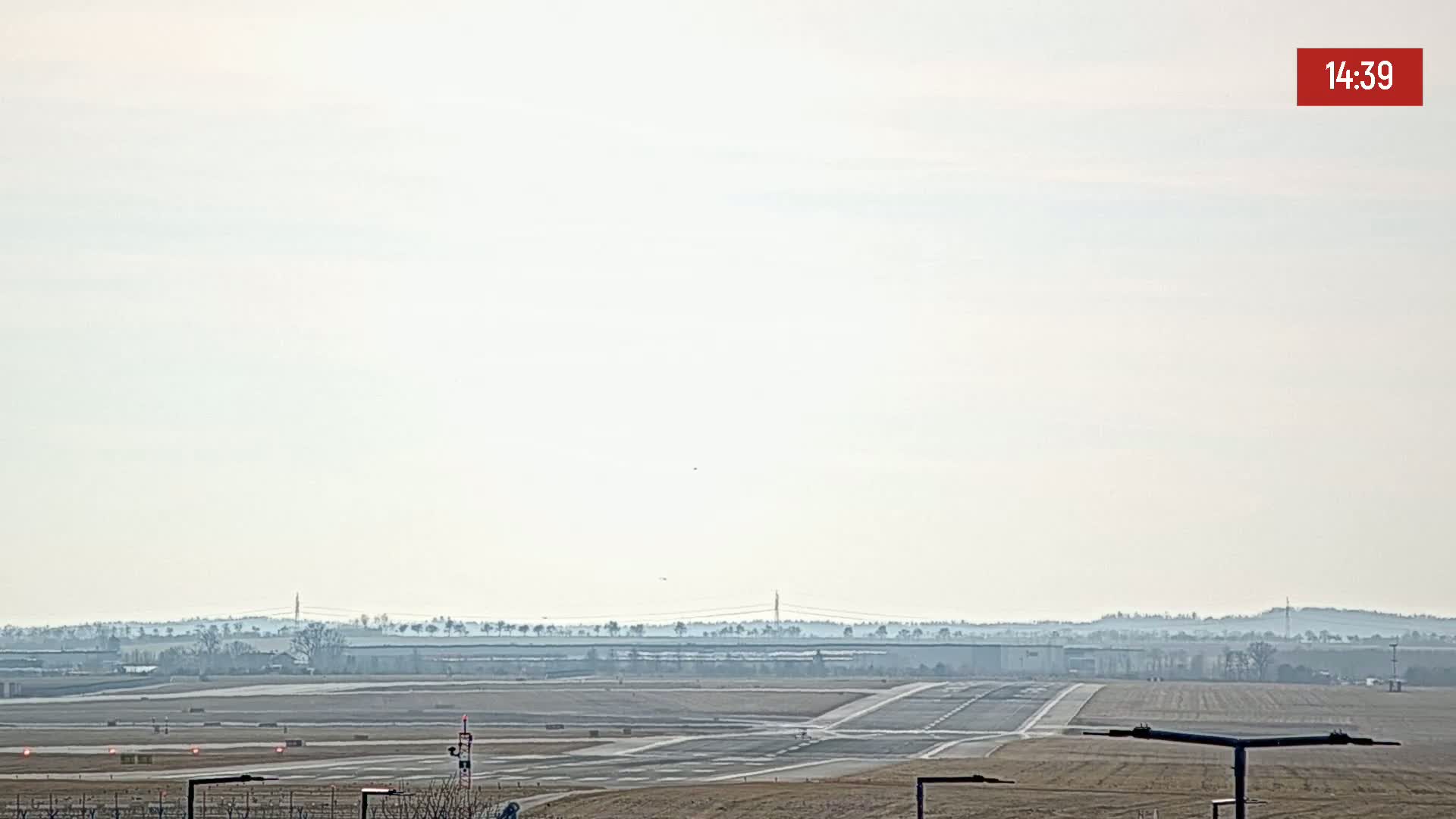 A hazy, light-colored sky overlooks an airport runway and taxiways, with a treeline visible in the distance.