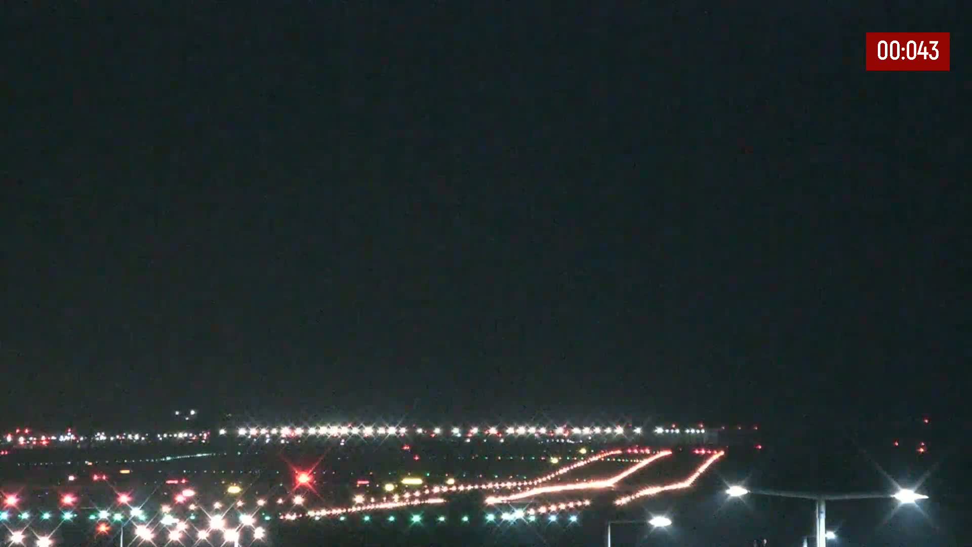 A nighttime long shot of an airport runway illuminated with various colored lights under a clear, dark sky.