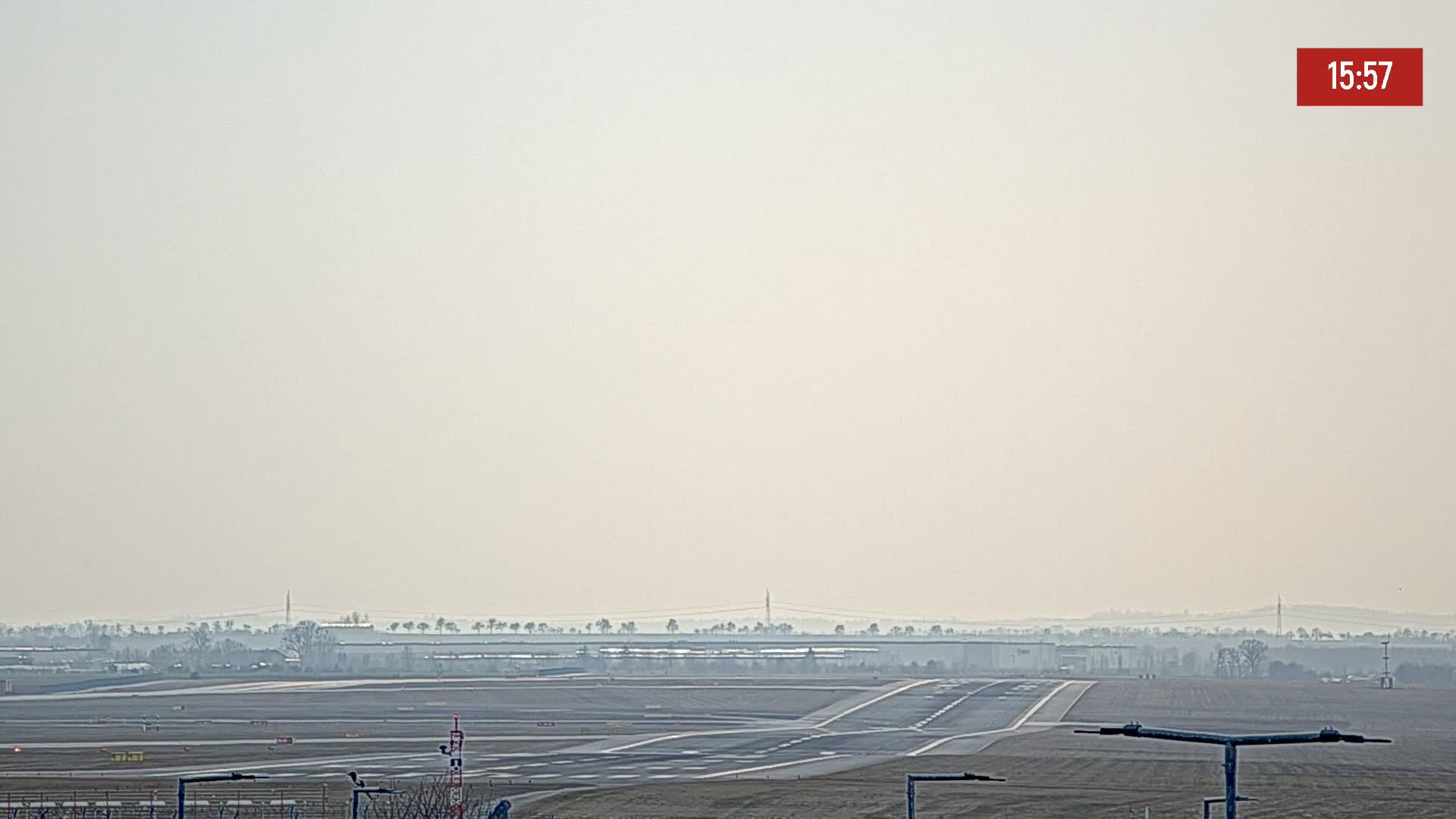 A hazy view of an airport runway and taxiways, with a distant horizon line of trees and buildings under a light, overcast sky.