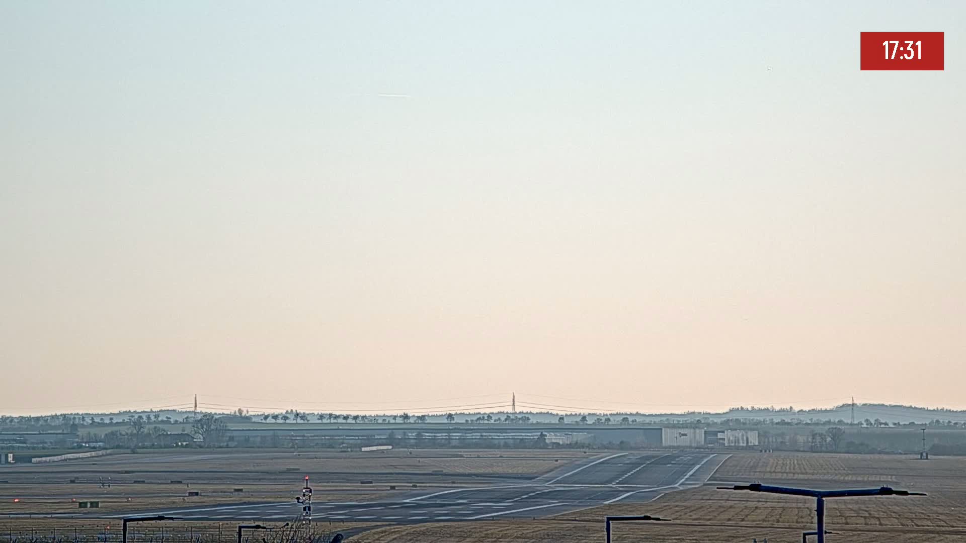 A runway at an airport on a hazy day extends toward a distant line of trees and buildings.