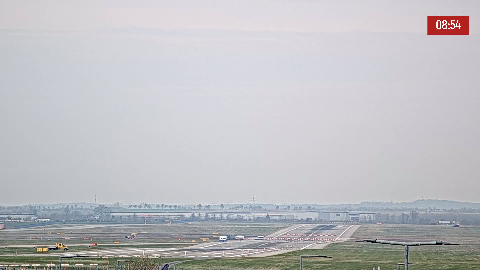 A hazy view of an airport runway with several vehicles, and some distant trees under an overcast sky.
