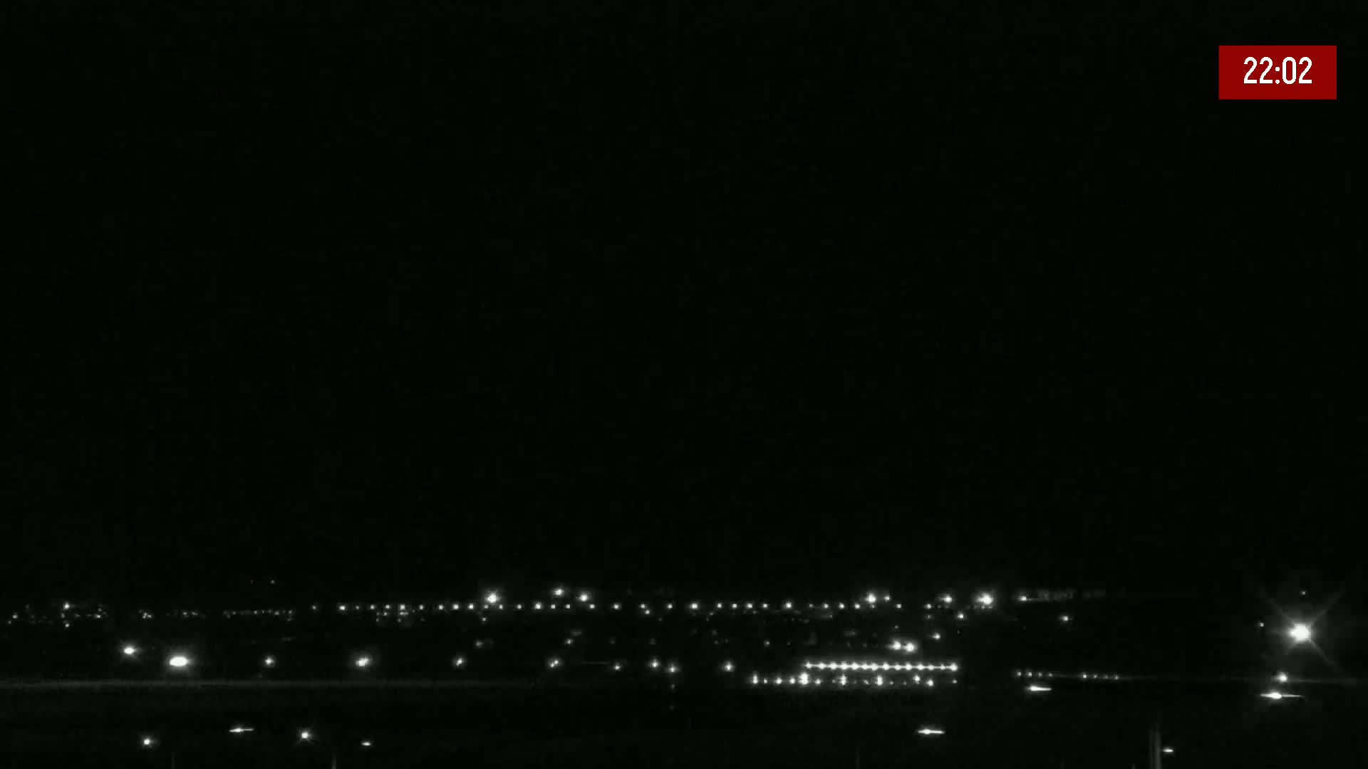 A nighttime long shot shows a distant town's lights spread across a dark landscape under a clear, black sky.