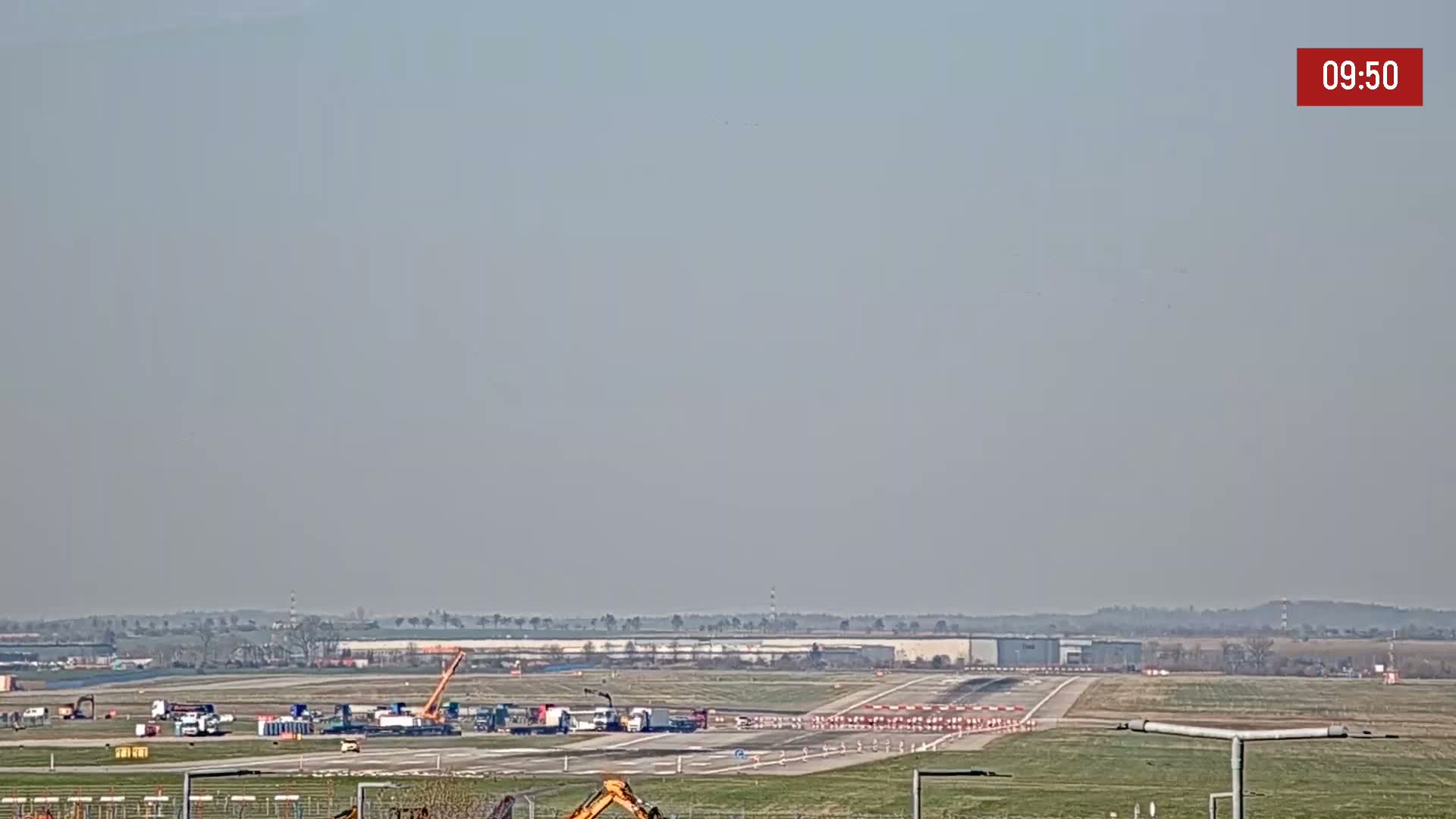 A hazy, light-colored sky overlooks an airport runway with several pieces of ground support equipment and construction vehicles present.