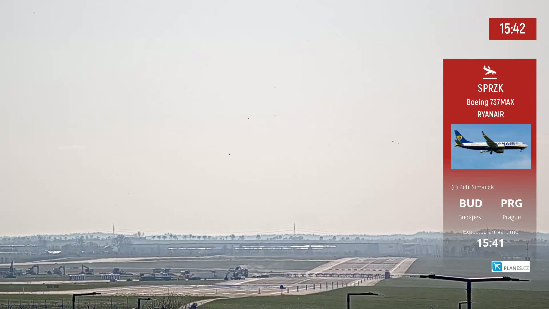A hazy view of an airport runway shows construction vehicles and a distant airplane approaching under a light, hazy sky.