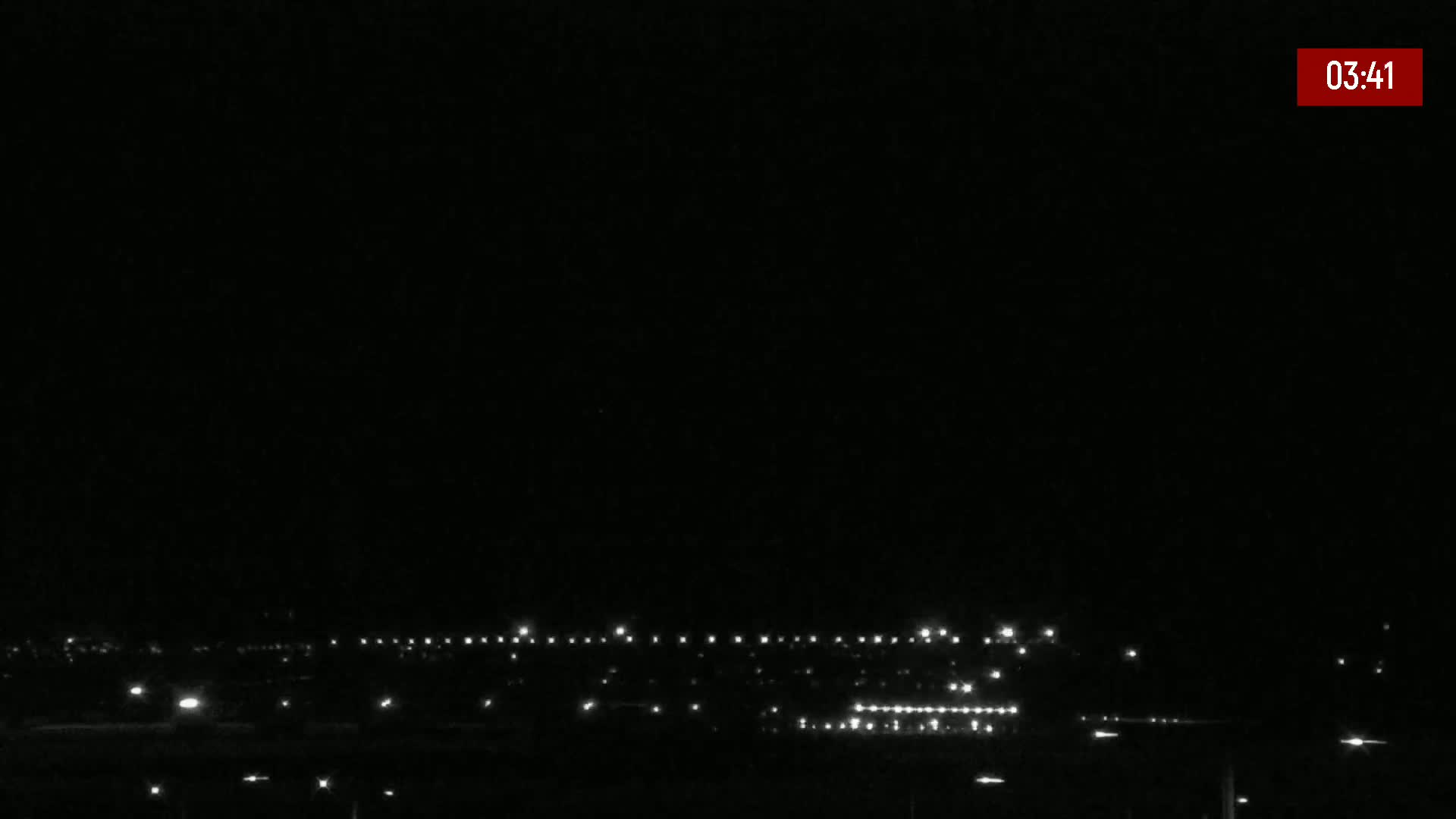 A nighttime long shot shows a distant line of lights from a town or city under a clear, dark sky.