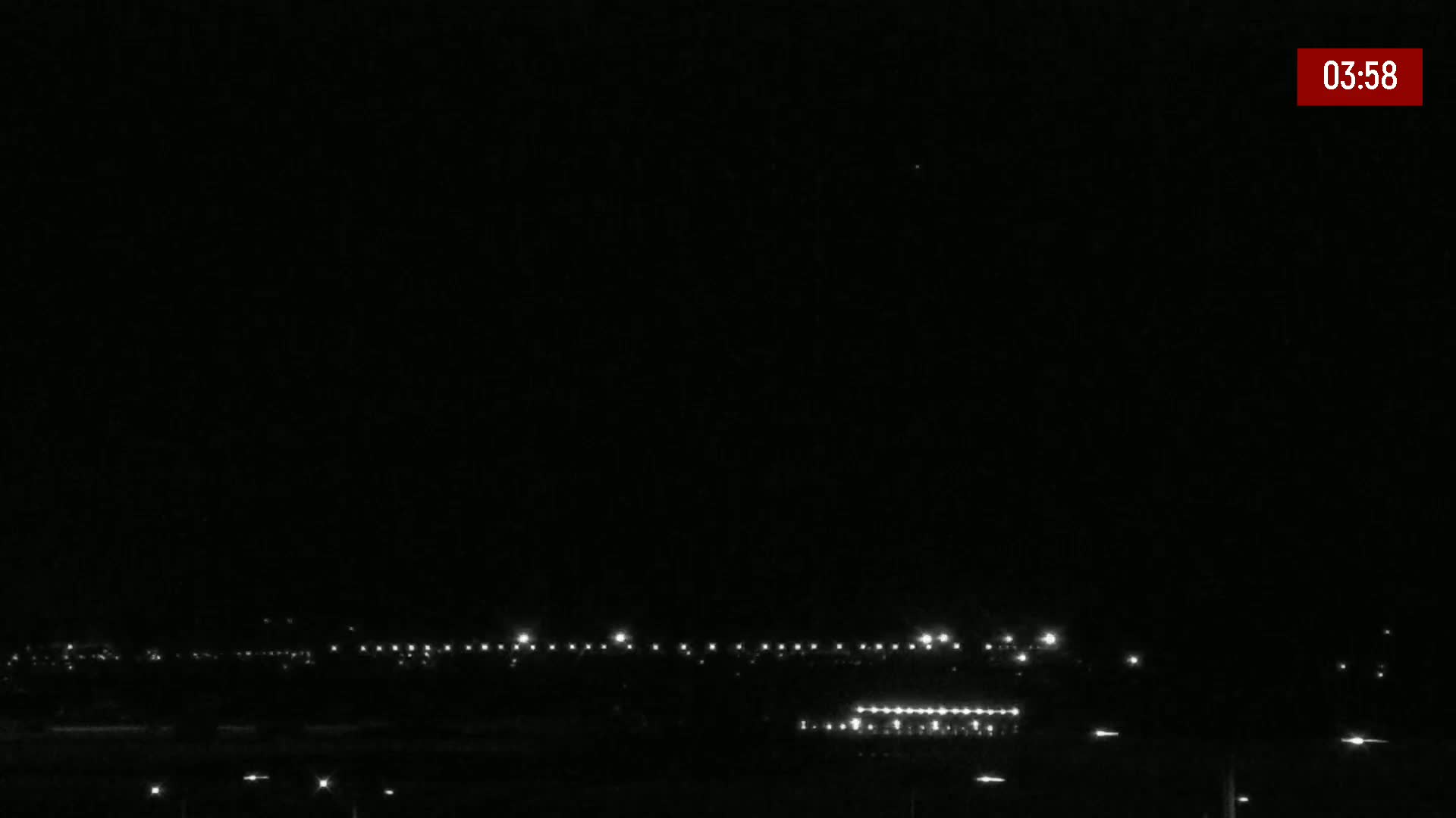 A nighttime long shot of a mostly dark landscape with a line of lights in the distance under a clear, dark sky.