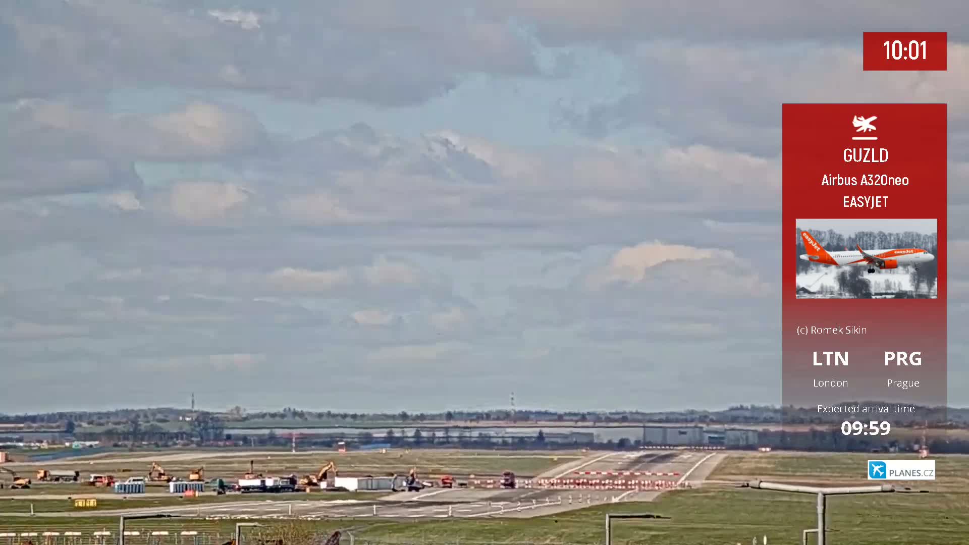 An airport runway with construction equipment is visible under a mostly cloudy sky.