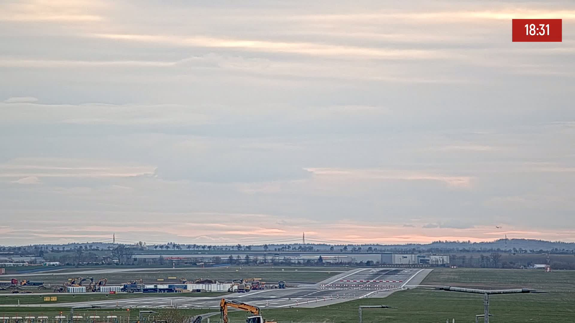 Under a pale, cloudy sky, an airport runway shows construction equipment and vehicles near the tarmac.