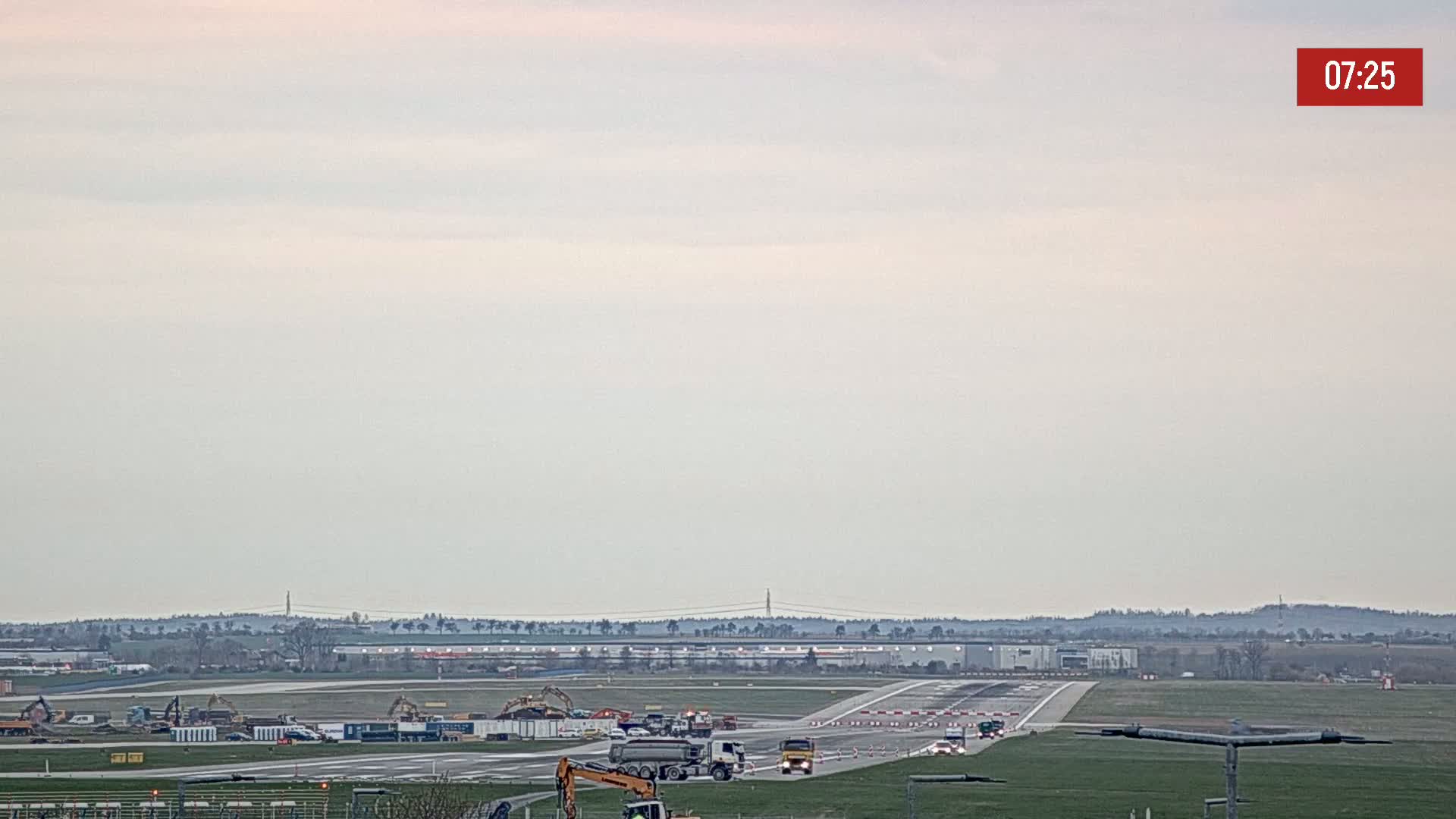 Under a pale, overcast sky, construction equipment and vehicles are visible on an airport runway.