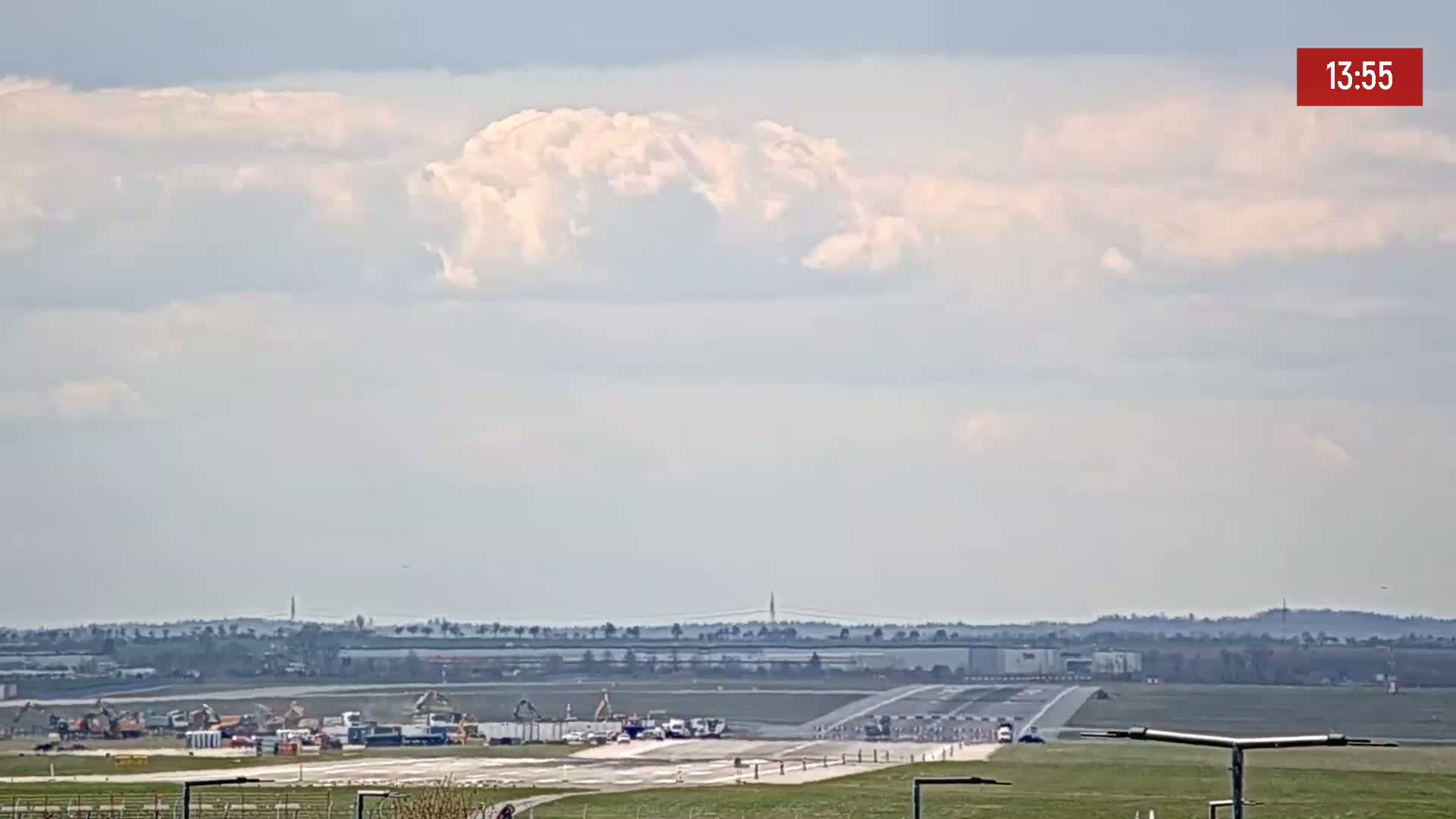A mostly cloudy sky overlooks an airport runway with several vehicles and construction equipment visible on and near the tarmac.