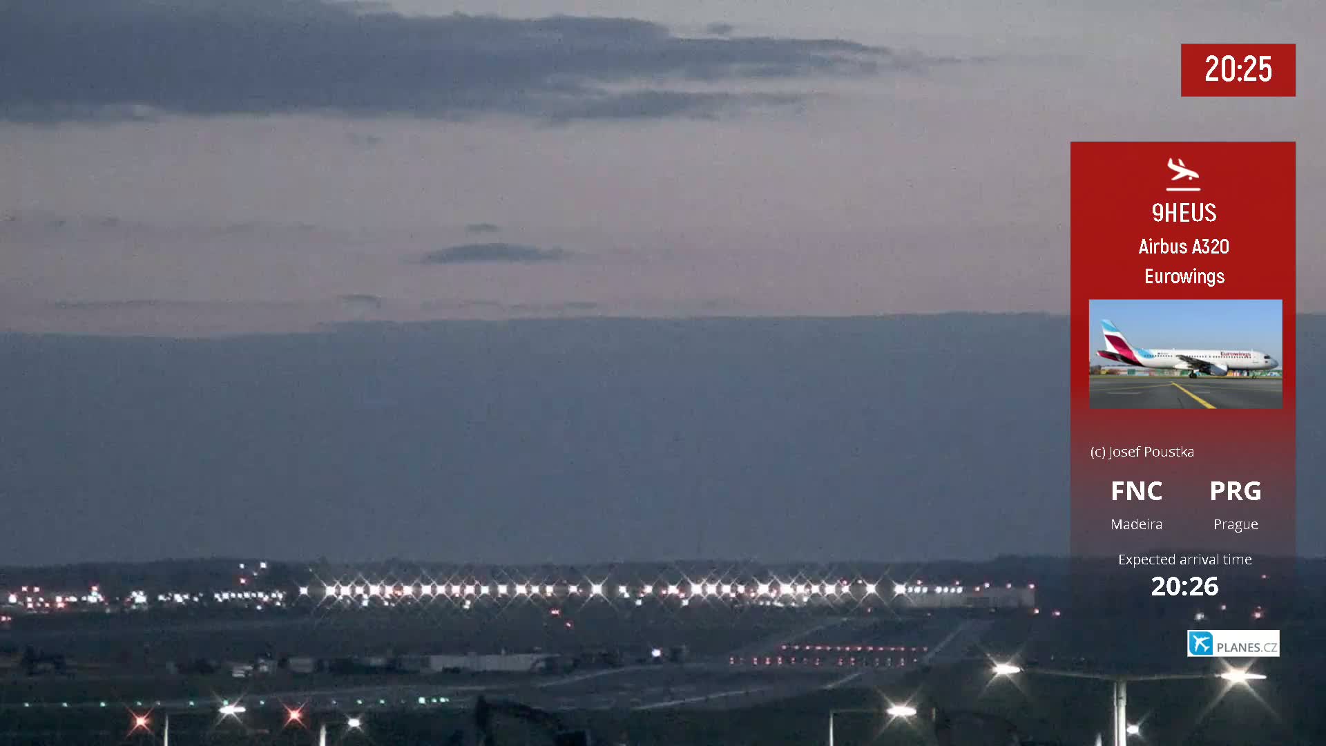 An airport runway at dusk, showing lights illuminating the tarmac and a plane approaching under a mostly cloudy sky.