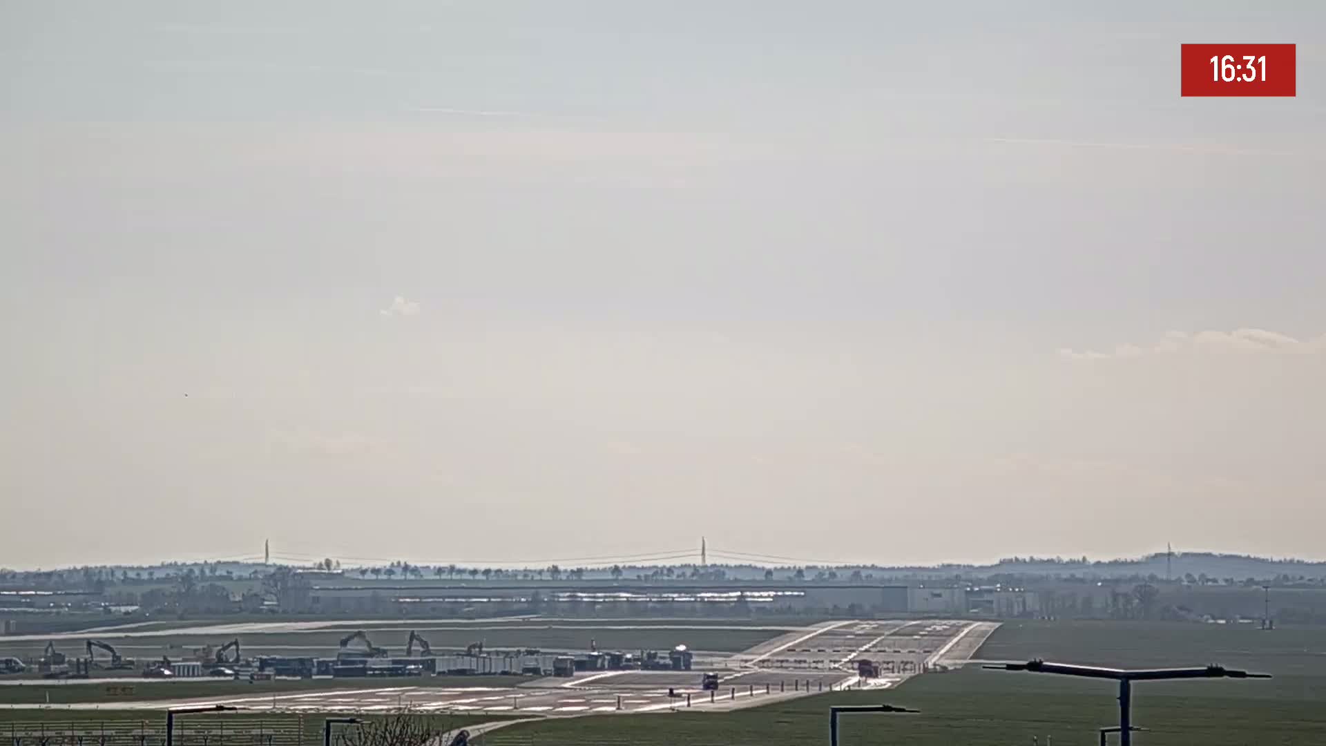 A long runway at an airport is visible under a hazy sky with a few scattered clouds, and construction equipment is seen near the tarmac.