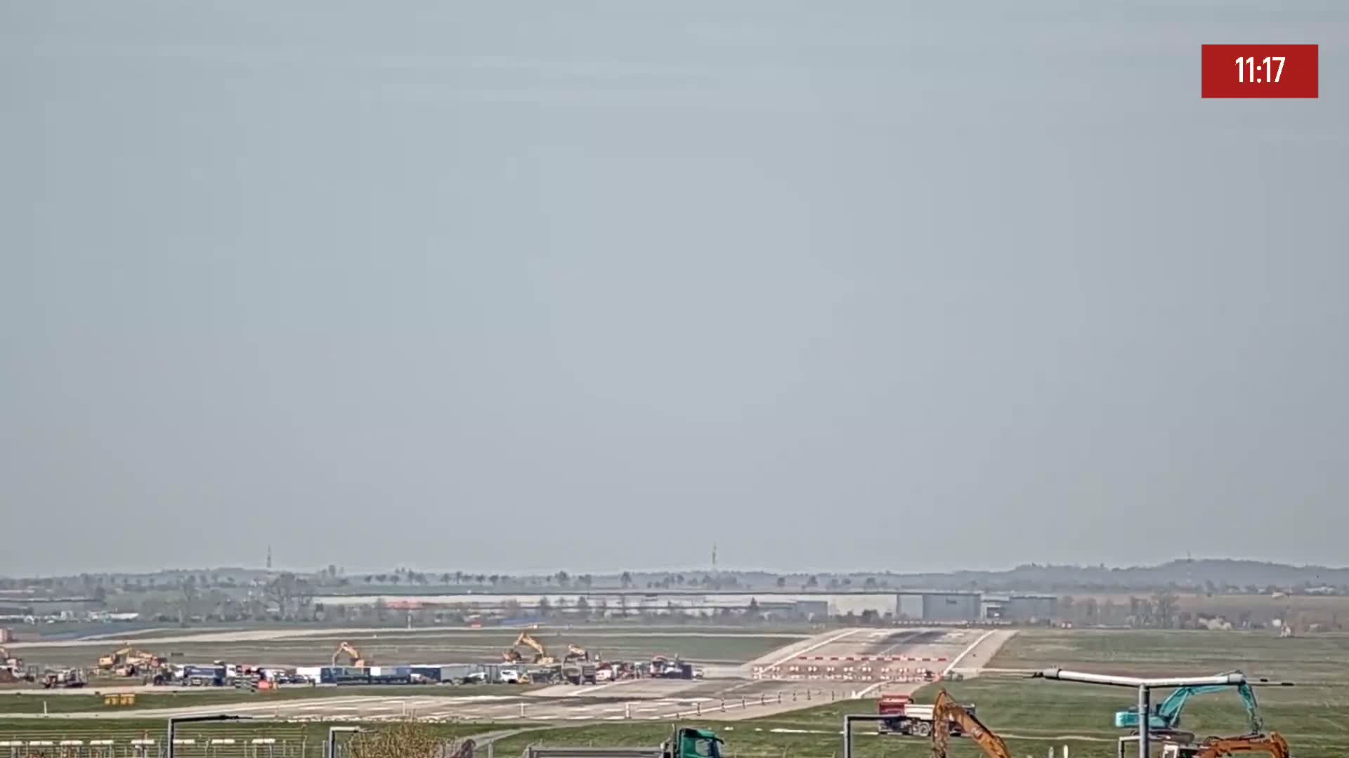 Under a hazy sky, construction equipment and vehicles are situated on an airfield runway.