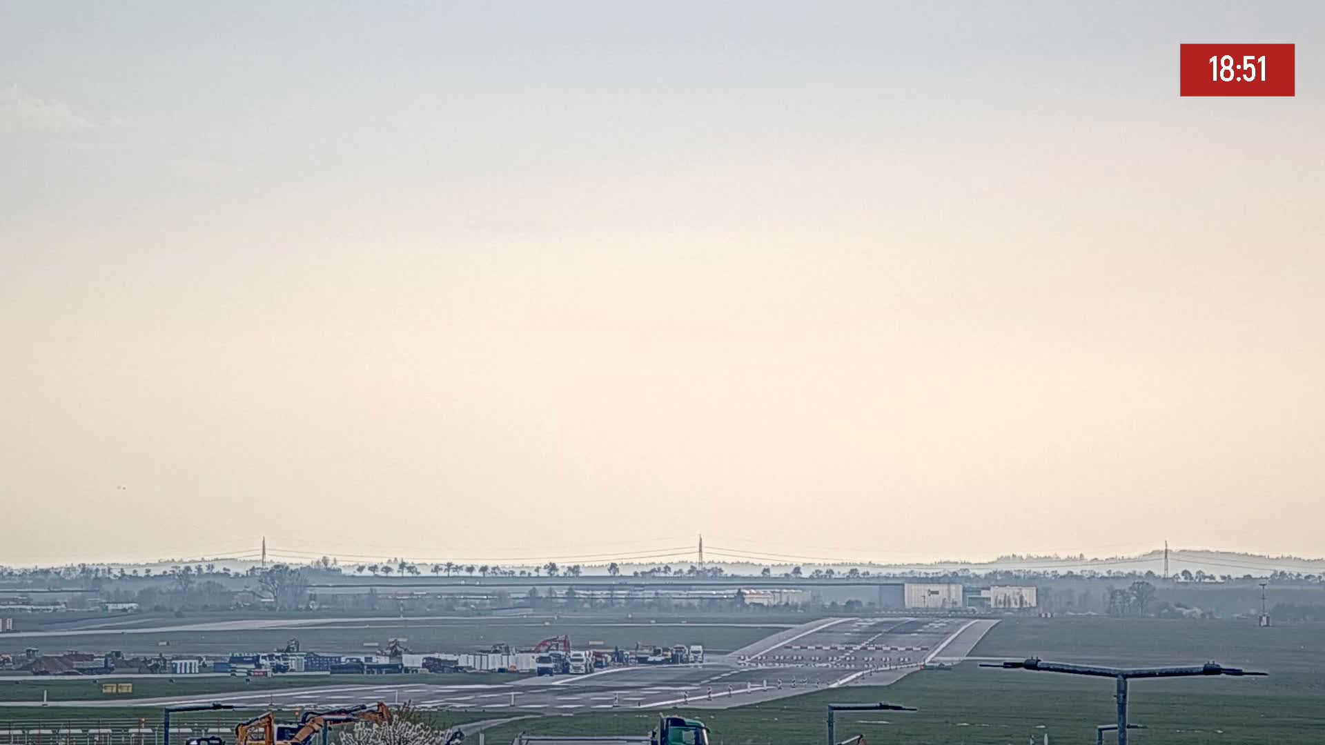 A hazy, light-colored sky overlooks an airport runway with several pieces of construction equipment and vehicles present.