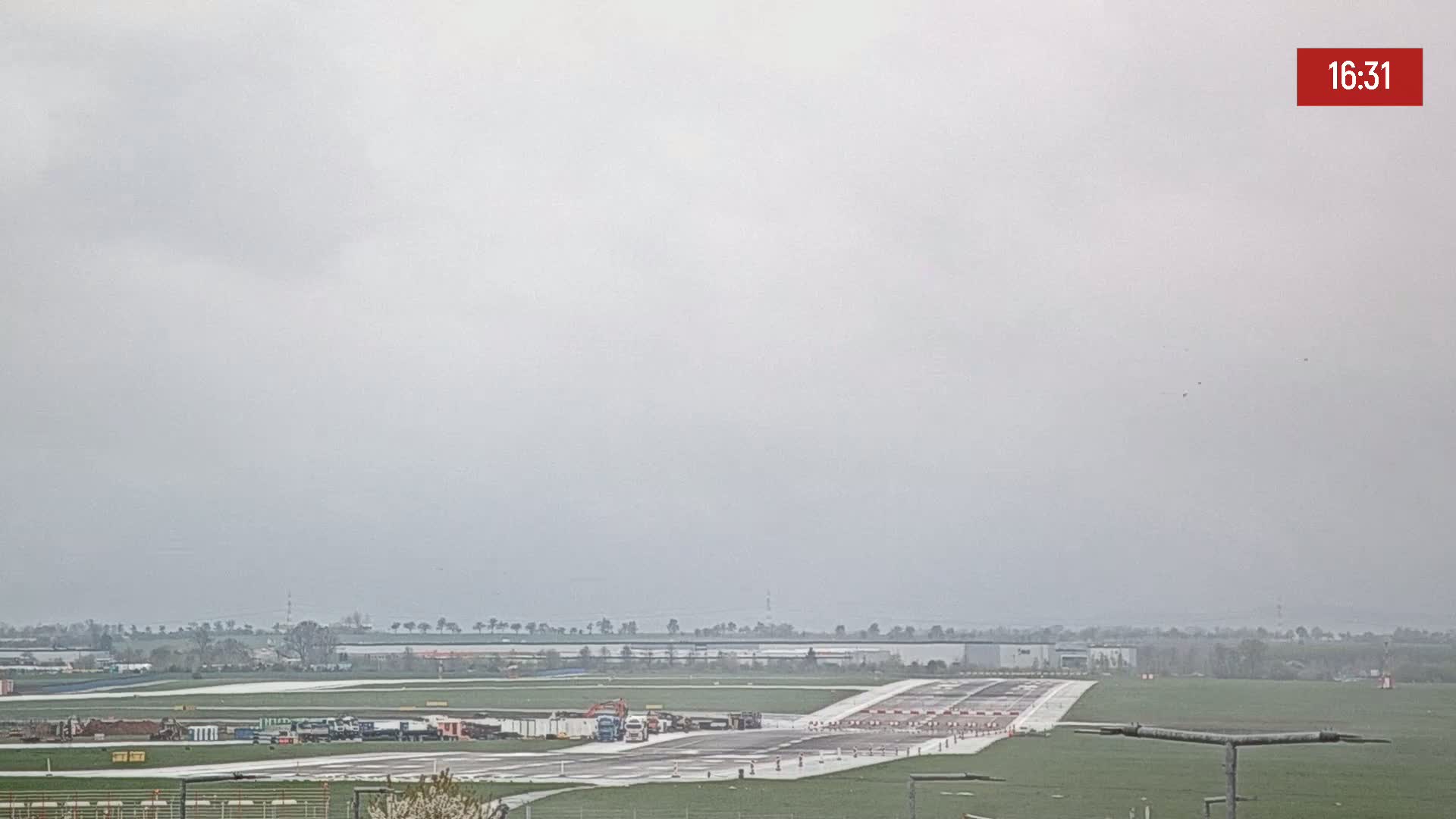 Under an overcast sky, several vehicles are parked on a runway near a flat expanse of land with sparse trees and buildings in the distance.