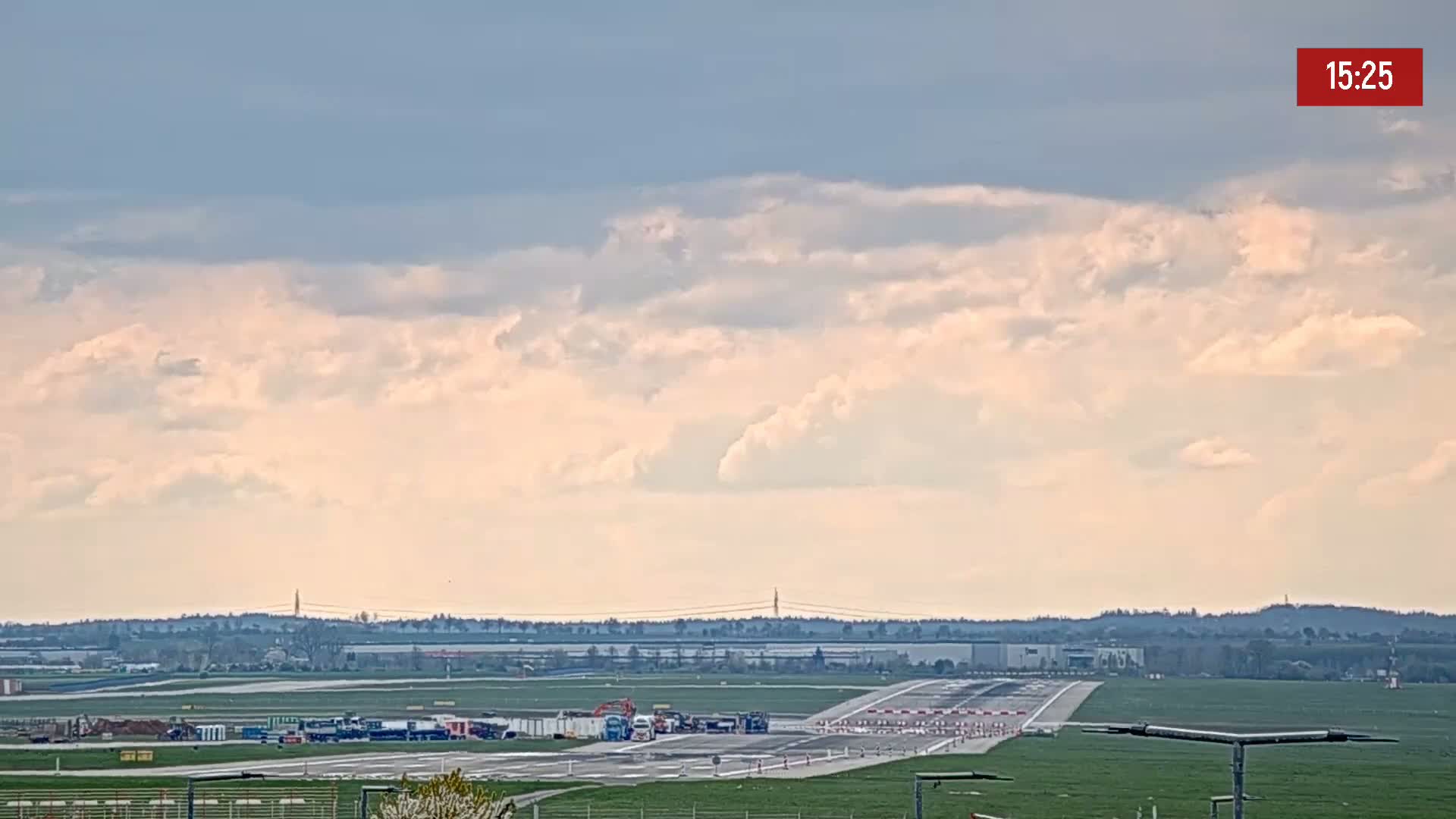 A partly cloudy sky overlooks an airport runway with several vehicles and equipment positioned near the end of the tarmac.
