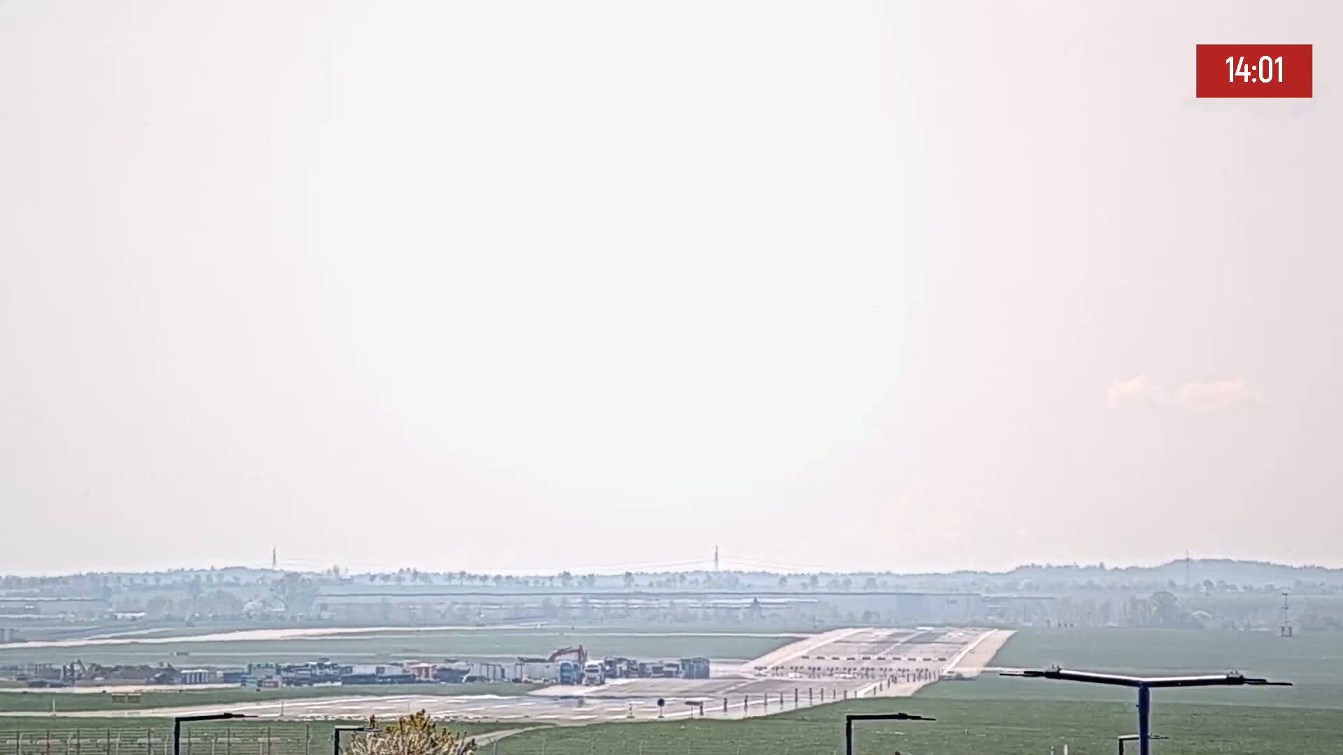 A hazy view of an airport runway with several vehicles parked near it and a treeline in the background.
