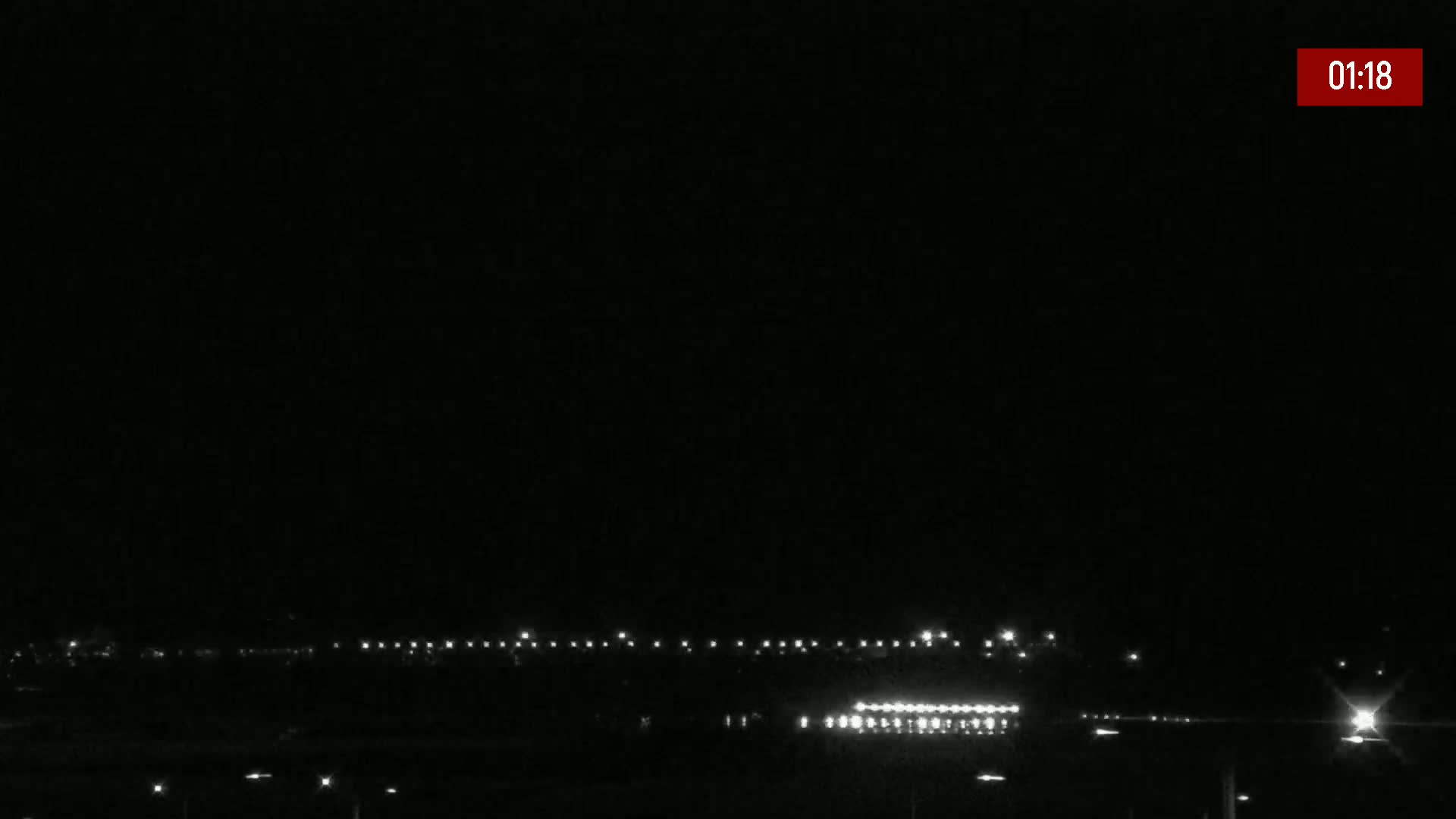 A nighttime, dark scene shows a distant line of lights, likely from a town or airport runway, under a clear, black sky.