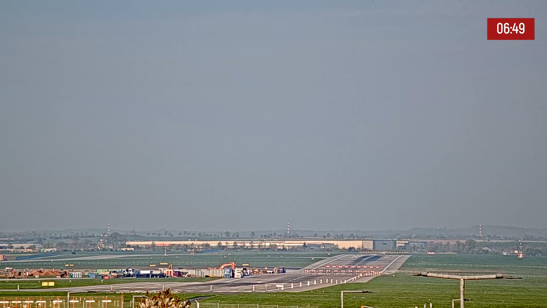 A hazy view of an airport runway with construction equipment and vehicles visible on the tarmac under a clear, light gray sky.