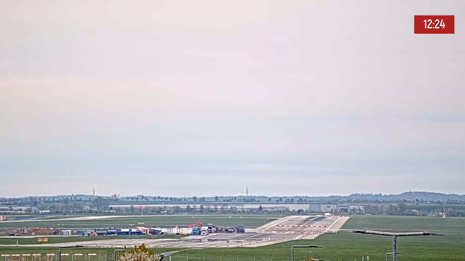A long runway at an airport is visible under an overcast sky, with several vehicles near the end of the runway and a distant horizon.