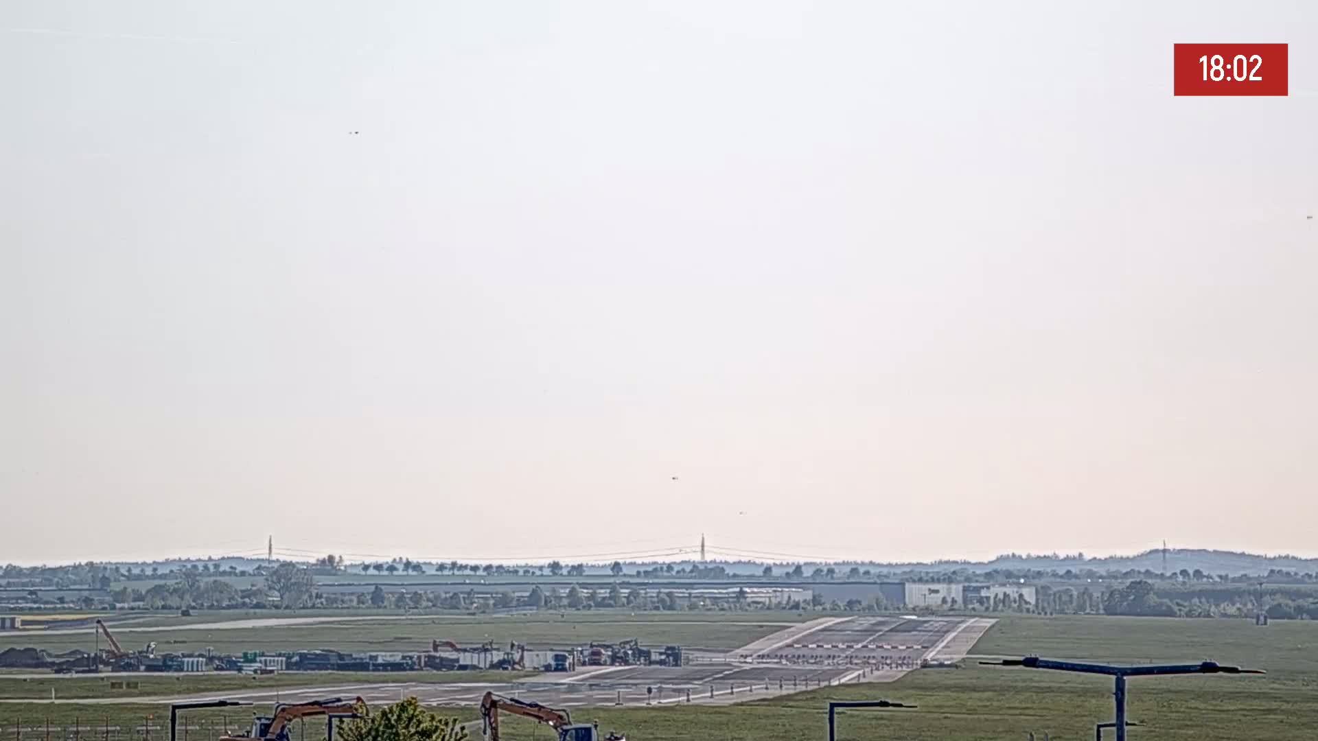 A hazy, light-colored sky hangs over an airport runway undergoing construction, with several pieces of heavy machinery visible.