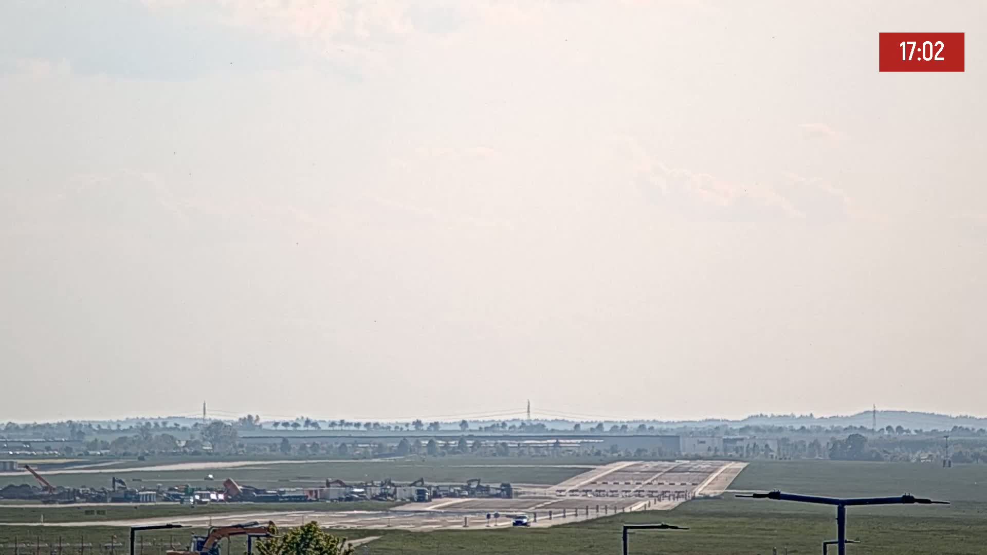 A hazy view of an airport runway with construction equipment and vehicles visible under a mostly cloudy sky.