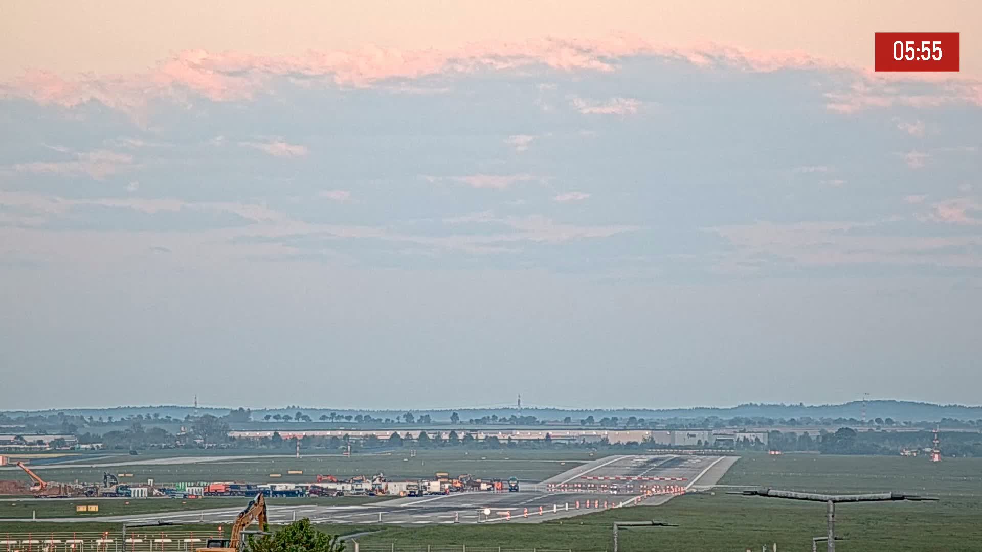 Under a pale pink and blue sky, construction equipment is visible on an airport runway.