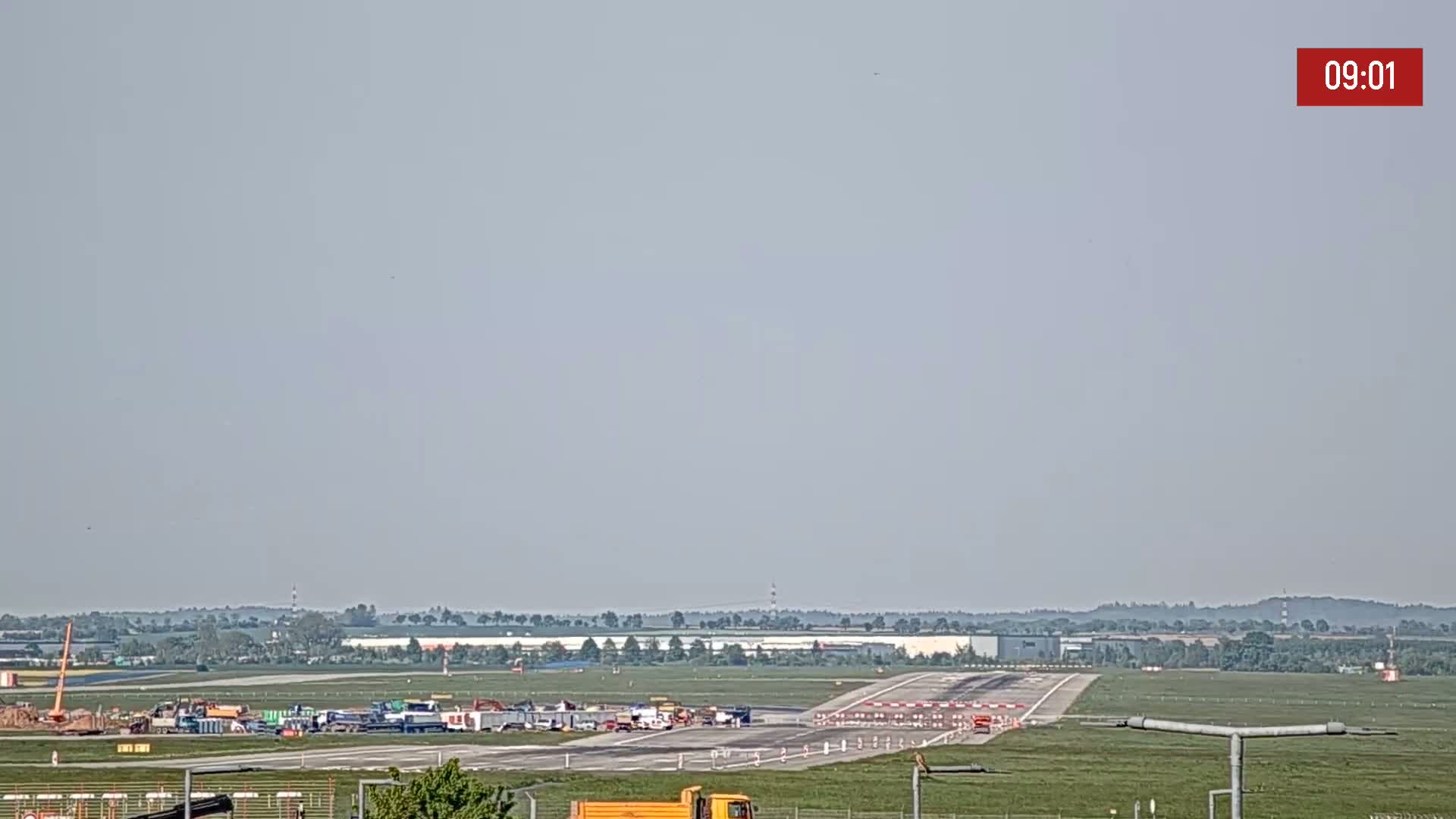 A mostly clear, hazy day reveals an airport runway with construction equipment and vehicles parked near the threshold.