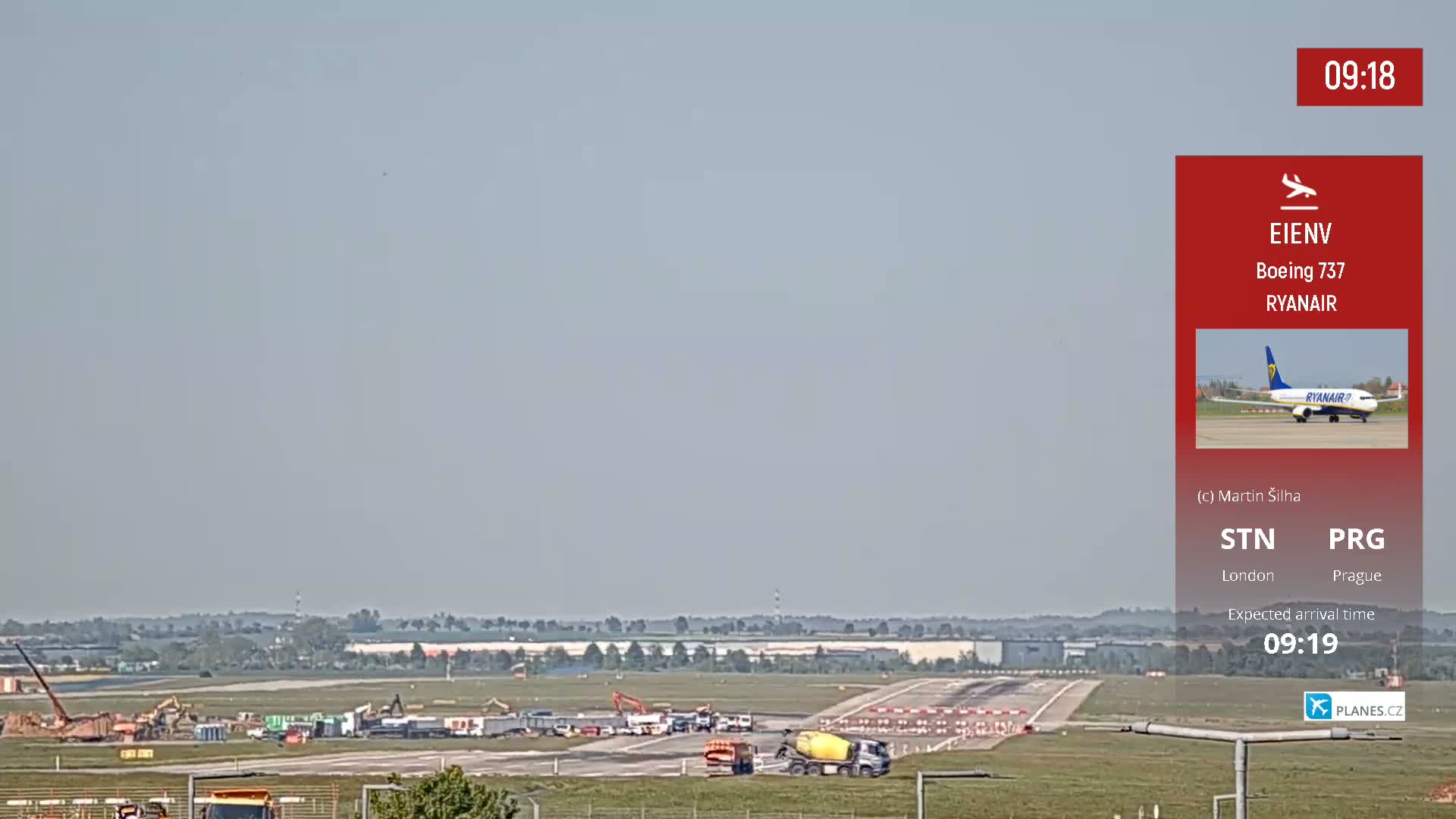 Under a hazy sky, a long runway shows construction activity with several pieces of heavy machinery and a cement mixer, while a distant airplane is visible on the tarmac.