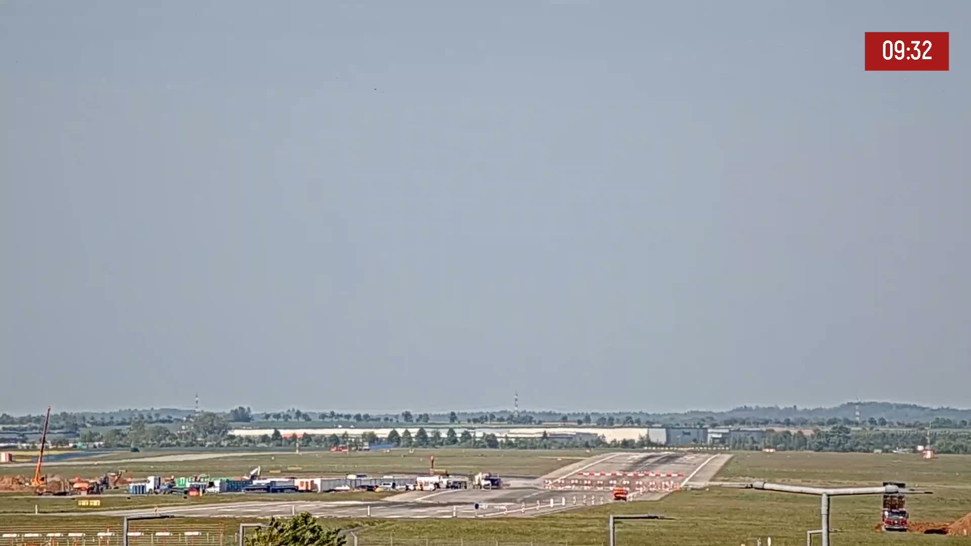Under a hazy sky, construction vehicles are visible on an airport runway surrounded by flat, grassy land.