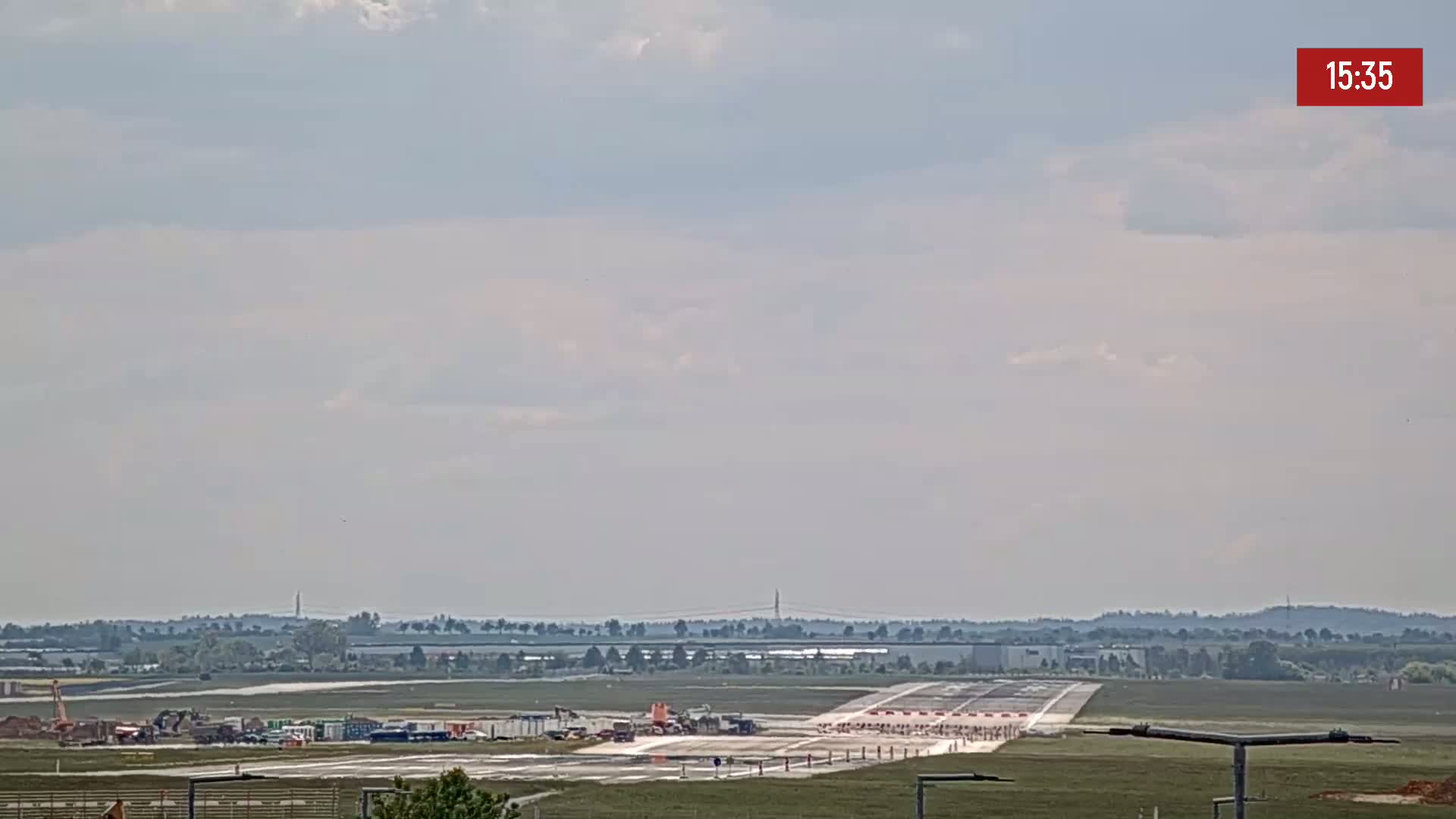 A long runway at an airport is visible under a partly cloudy sky, with construction equipment and workers present on the tarmac.