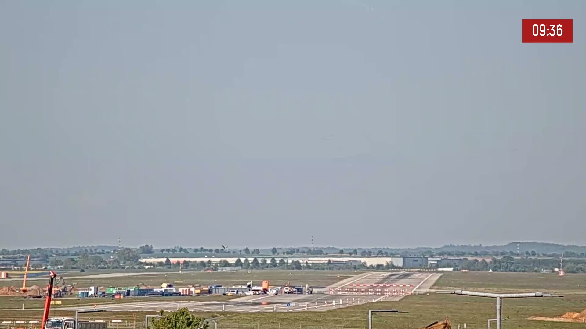 A long shot of an airport runway under hazy skies shows construction equipment and vehicles near the runway.