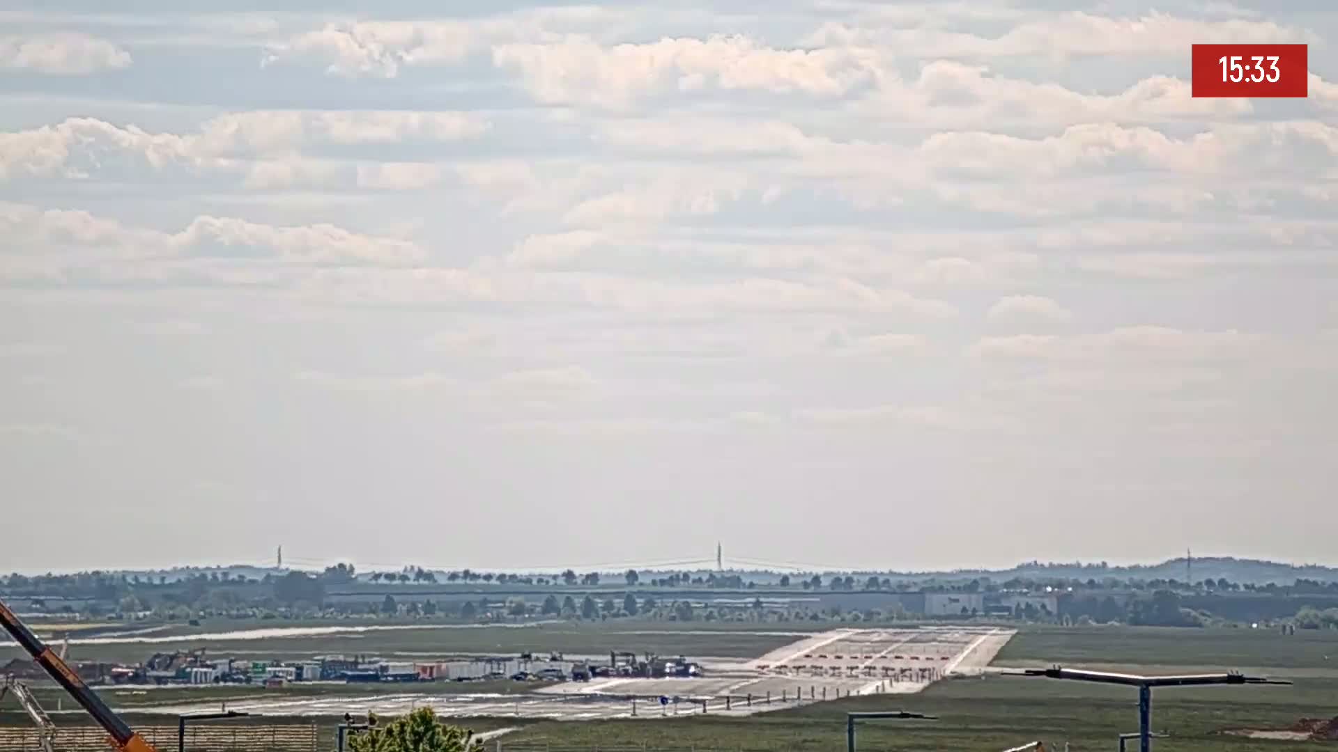 Under a partly cloudy sky, a long runway is visible with construction equipment and vehicles near it, along with a distant landscape of trees and buildings.
