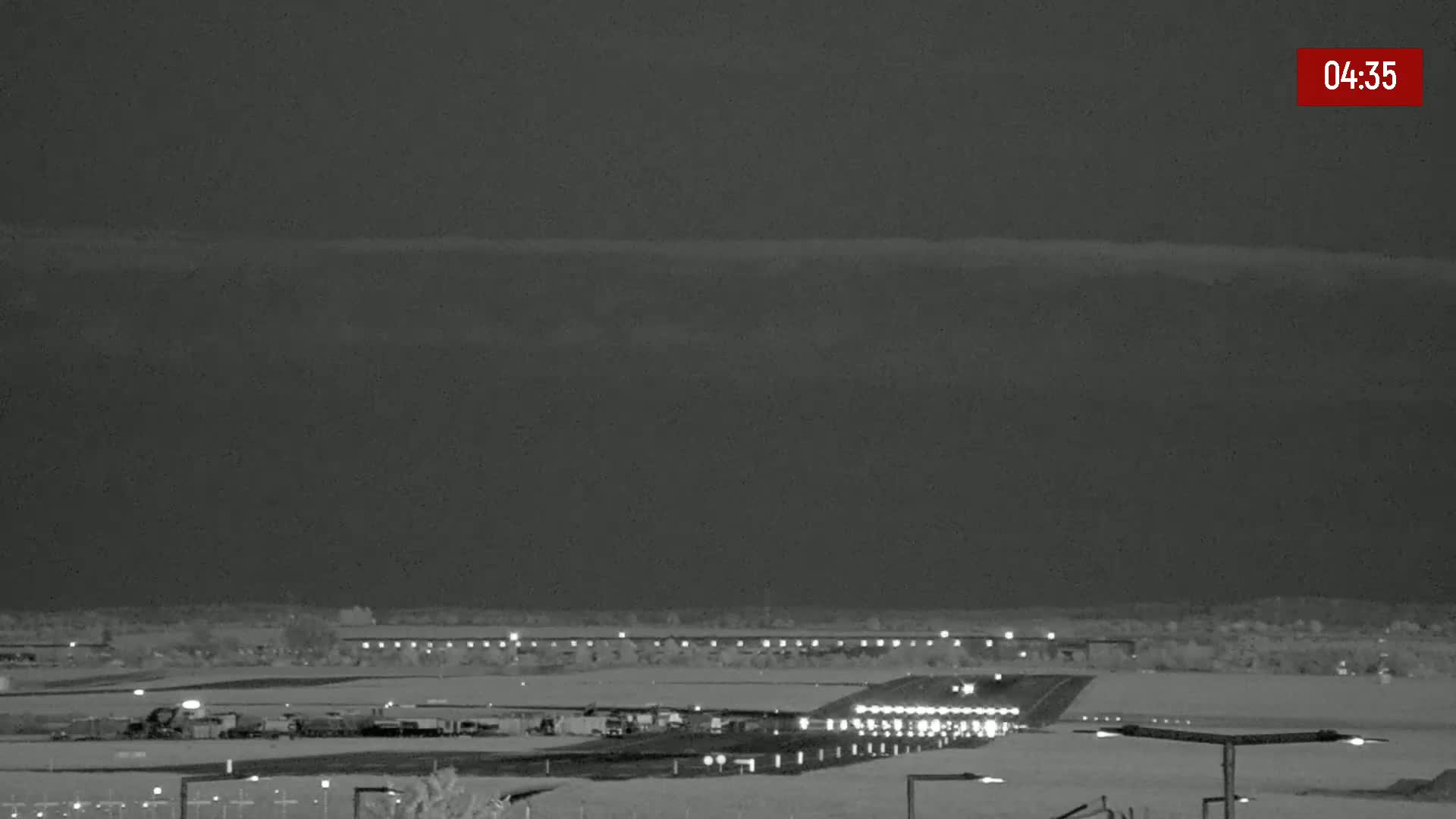 An airport runway is visible at night under a cloudy sky, with various vehicles and lights illuminating the tarmac.