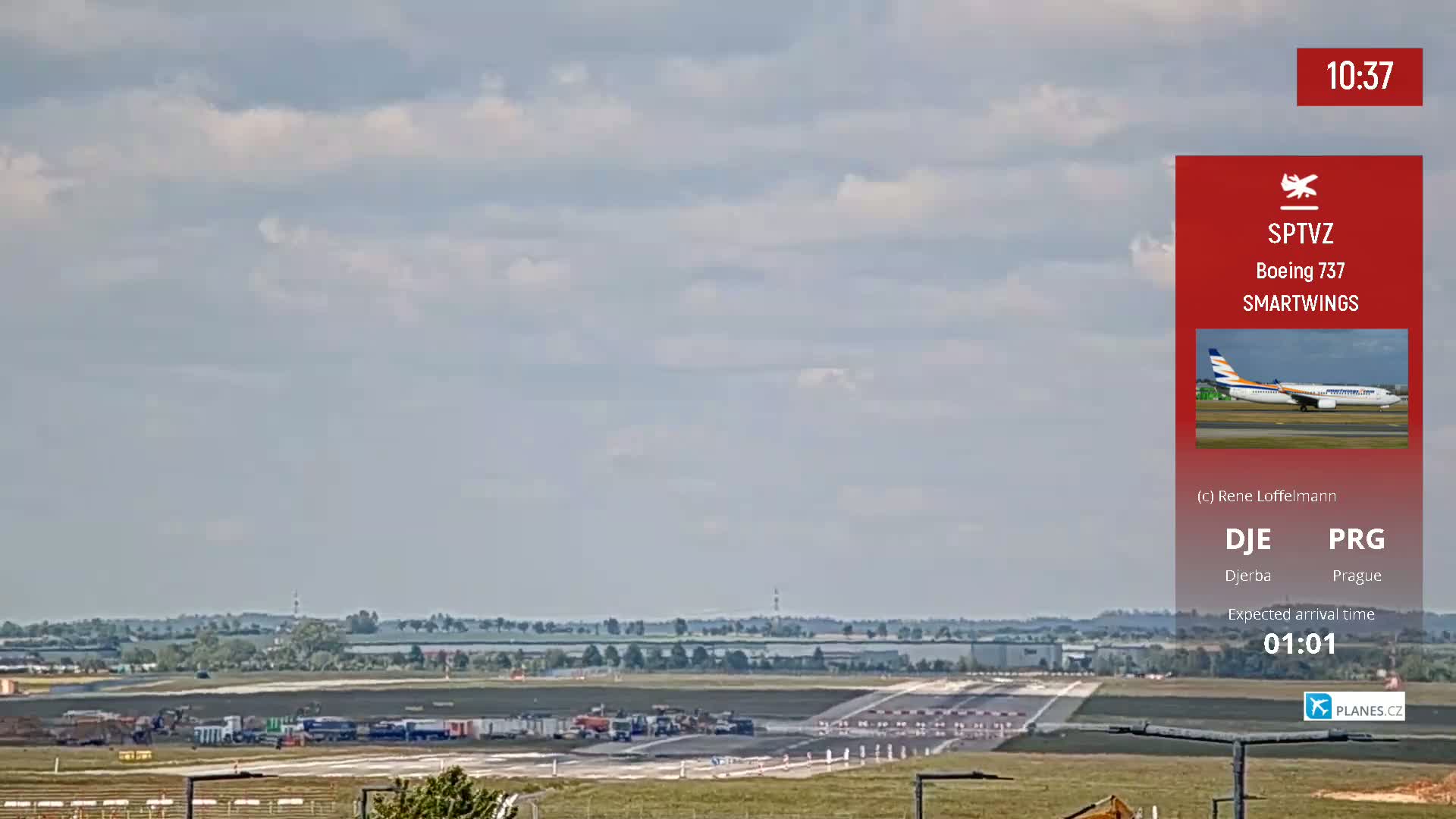 A mostly sunny day at an airport shows a runway with several ground vehicles, and a distant landscape of flat green fields under a partly cloudy sky.