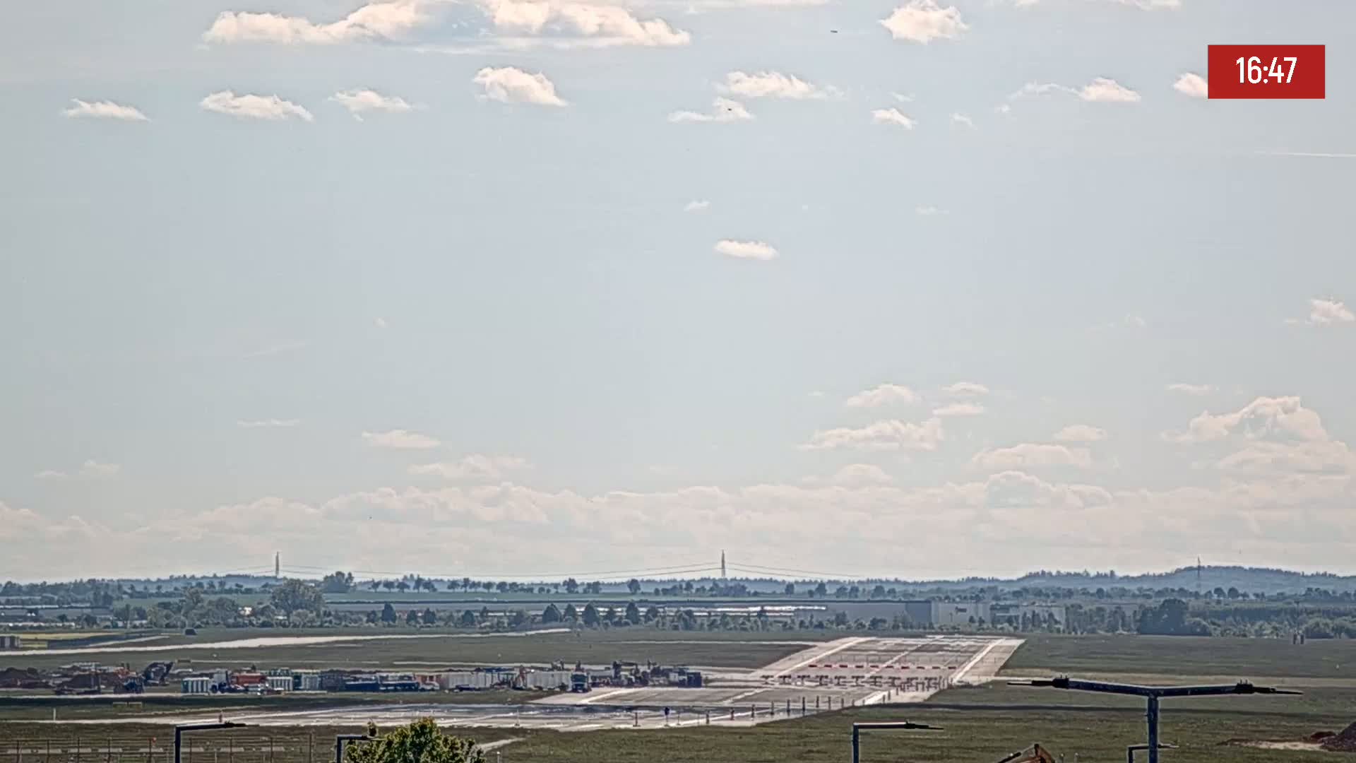 A mostly sunny day reveals an airport runway with construction equipment and vehicles near the terminal, surrounded by flat land and distant trees under a partly cloudy sky.