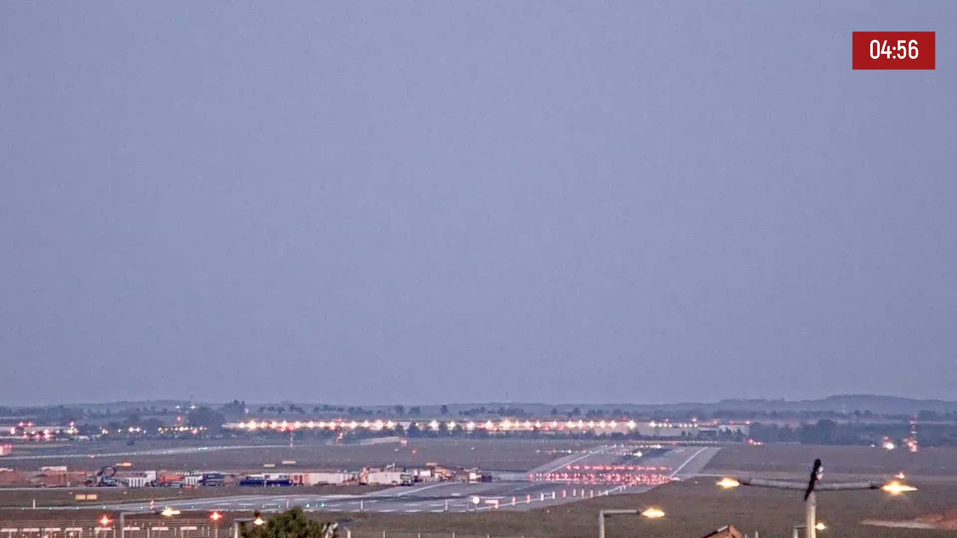 Under a twilight sky, an airport runway is shown with various vehicles and equipment present.